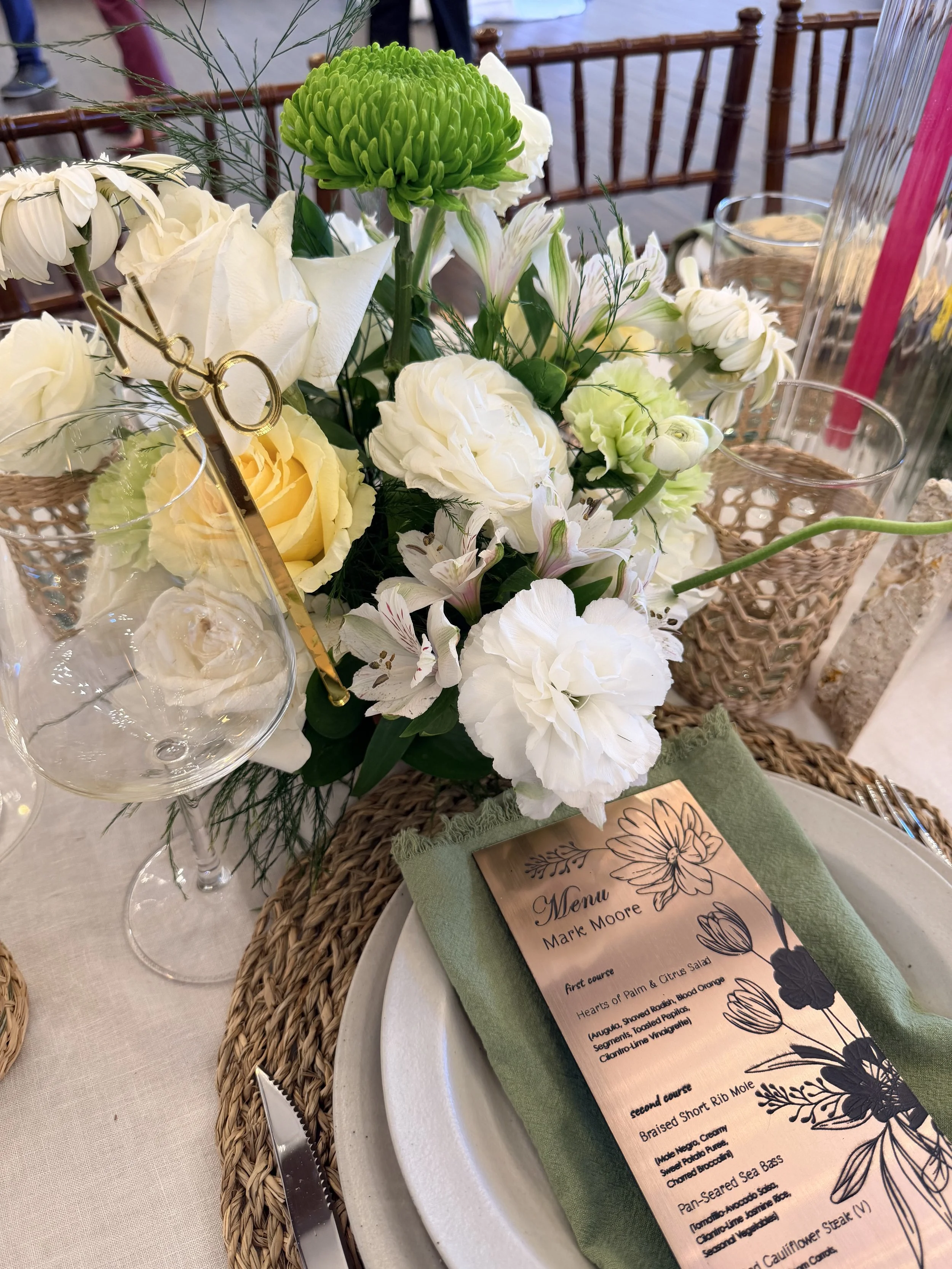 Close-up of a floral centerpiece with white and green flowers, a menu card, glassware, and cutlery on a woven placemat at a dining table.