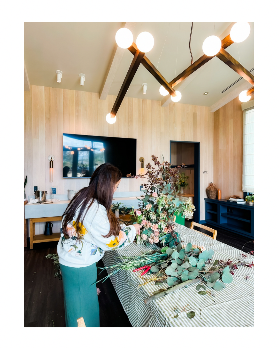 Woman arranging flowers on a table in a modern room with wood-paneled walls and a large flat-screen TV.