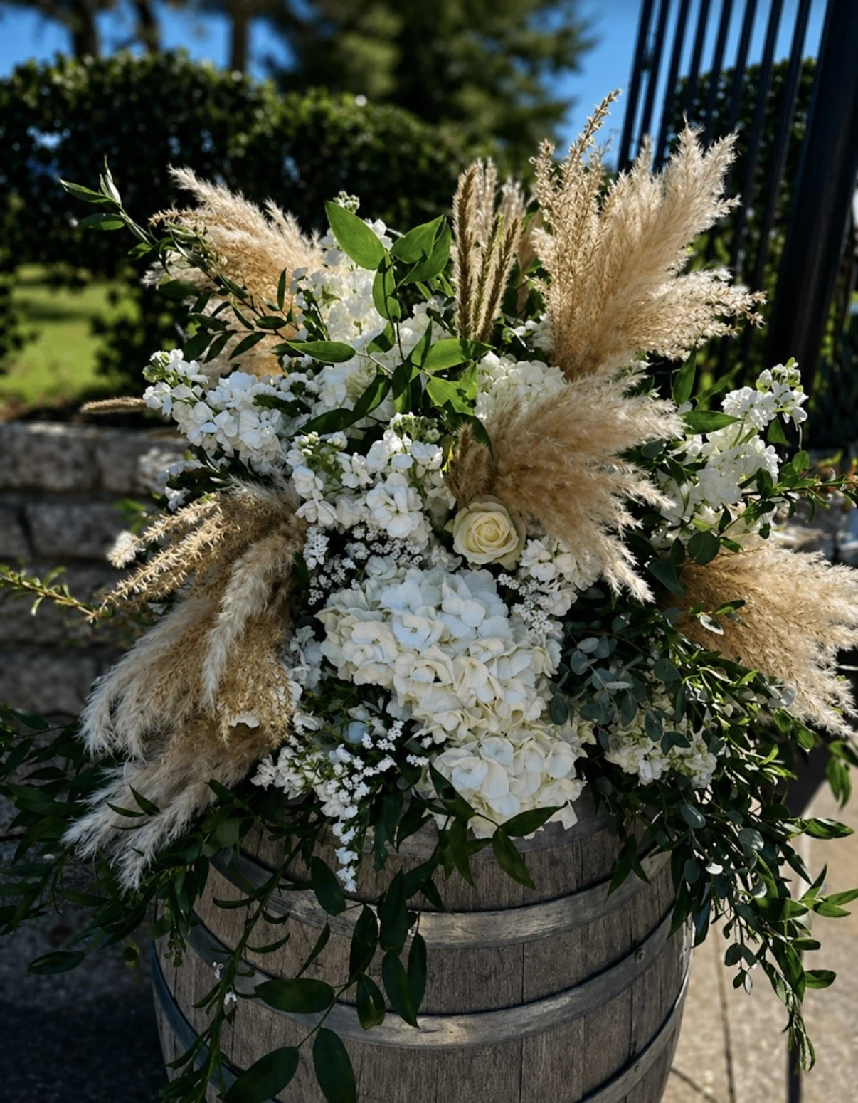 A floral arrangement with white flowers, greenery, and tall beige pampas grass in a wooden barrel planter outdoors.