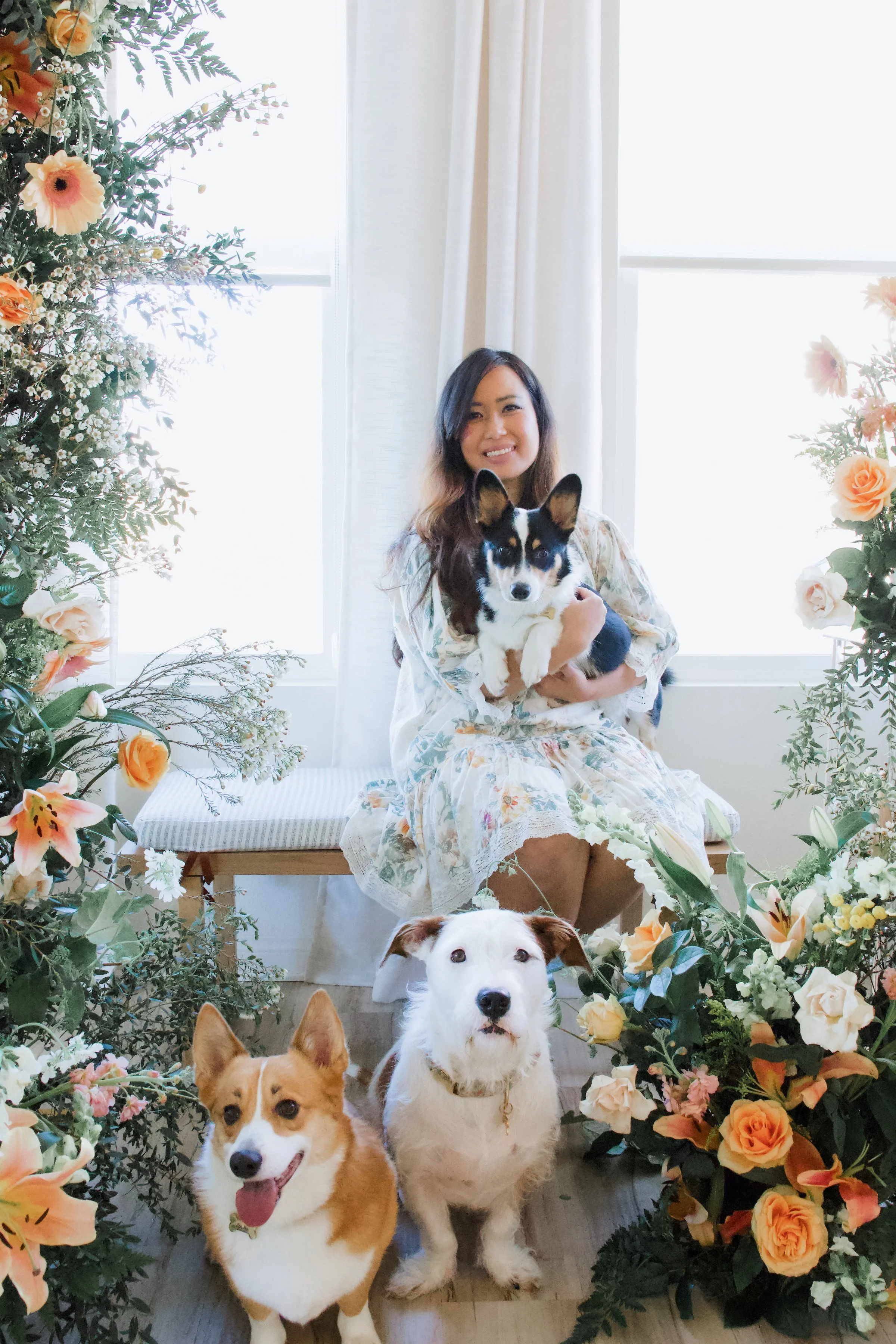 A woman in a floral dress sitting on a bench holding a black and white dog, surrounded by two other dogs and floral arrangements near windows with white curtains.