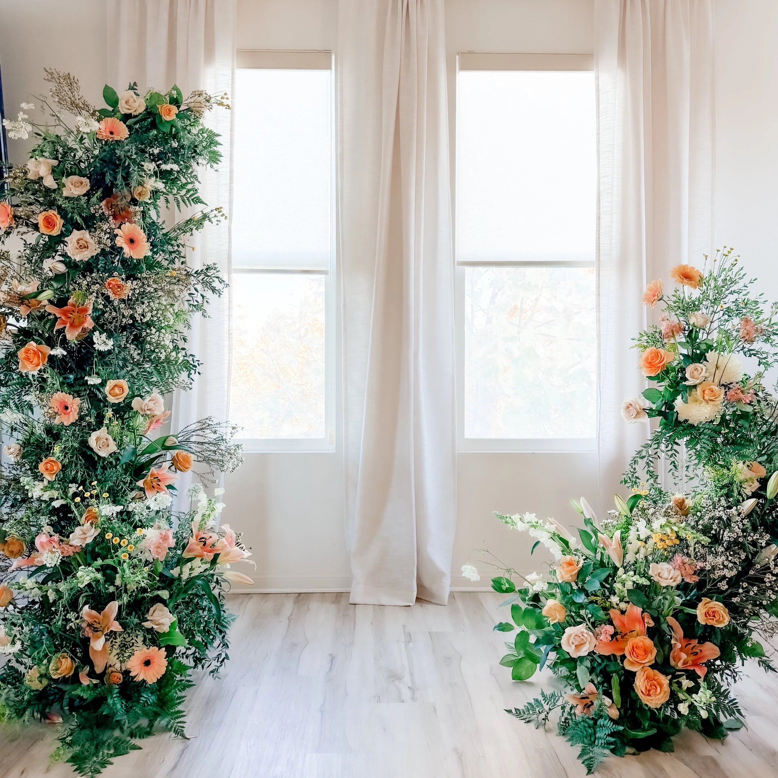 Two large floral arrangements with peach and white roses, lilies, and greenery placed on the floor near windows with white curtains.  San Diego Wedding Floral Arch Installations