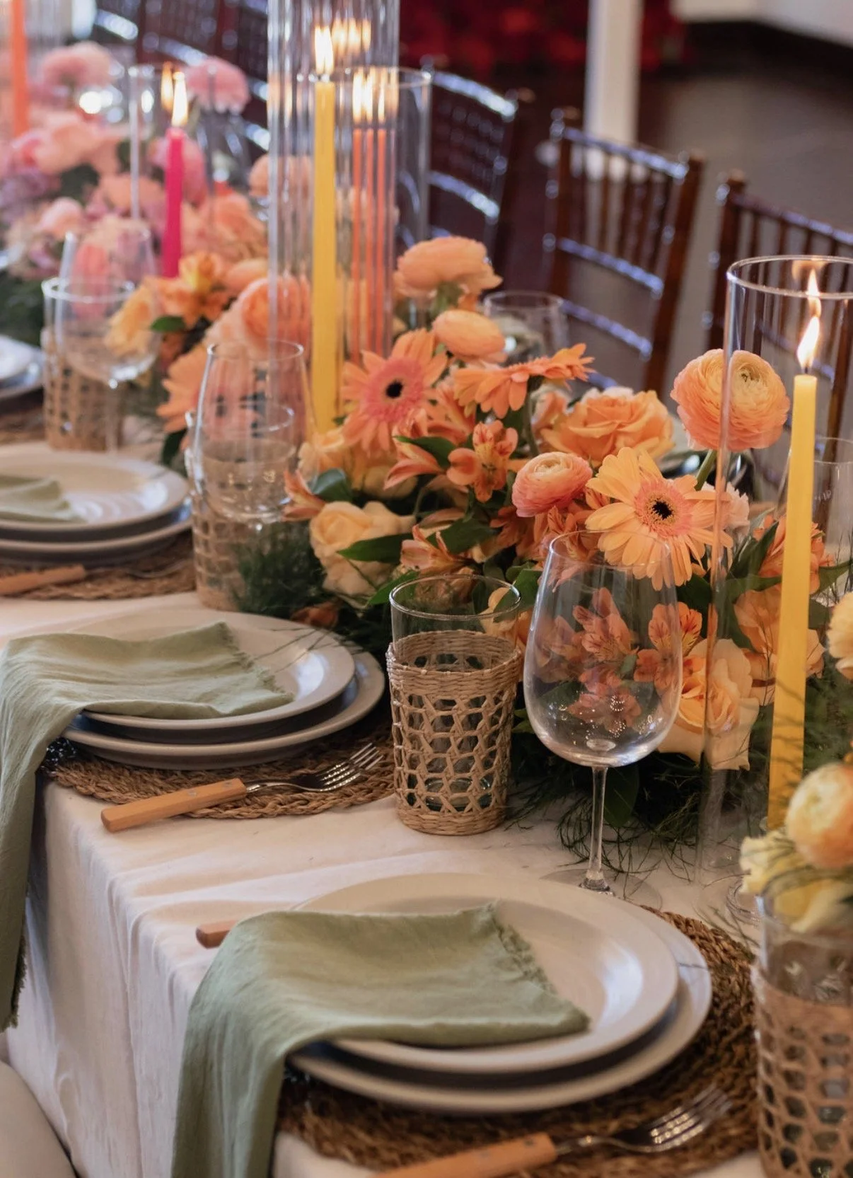 A decorated table with pink and peach flowers, tall yellow and pink candles in clear vases, wine glasses, plates with pale yellow napkins, and woven placemats, set for a celebration or event.