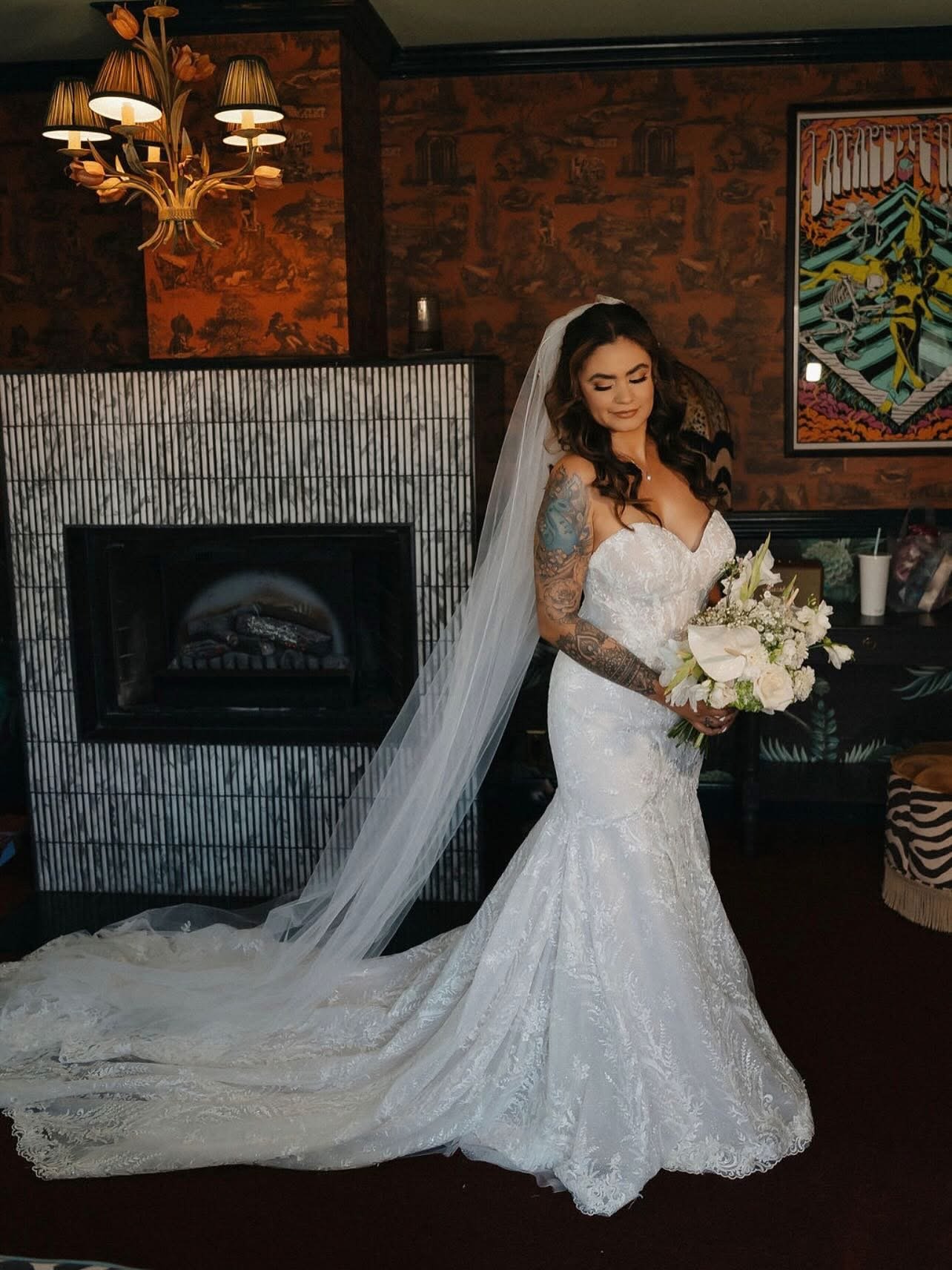 A bride with dark, curly hair wearing a white lace wedding dress with a long train and veil, holding a bouquet of white flowers, standing in a cozy room with a fireplace, decorative wall, and modern art.