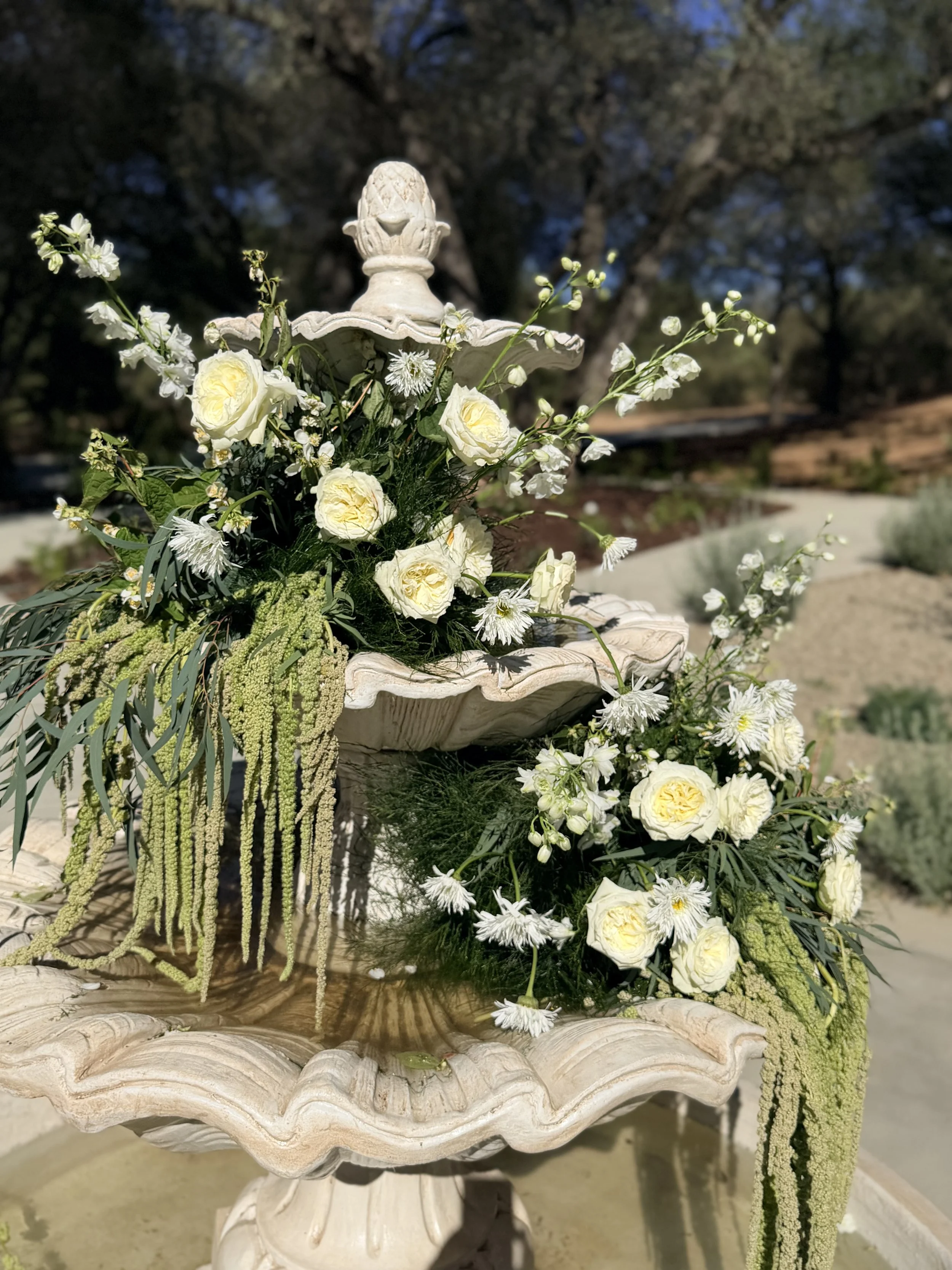 A white decorative fountain decorated with white roses, white daisies, and hanging green amaranthus flowers, set outdoors with trees in the background.