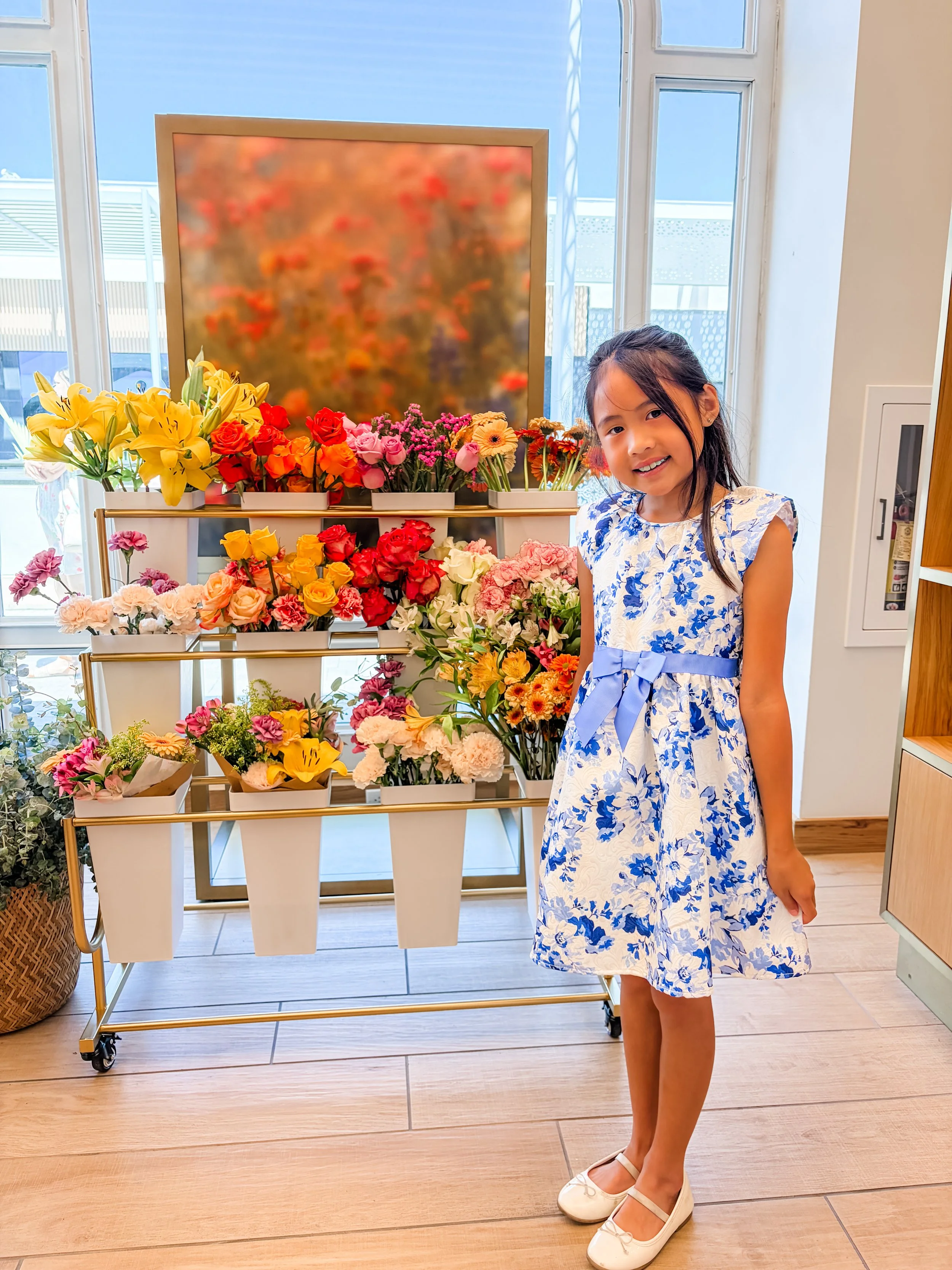 A young girl smiling in a floral blue and white dress stands in front of a display of colorful flowers inside a building with large windows.
