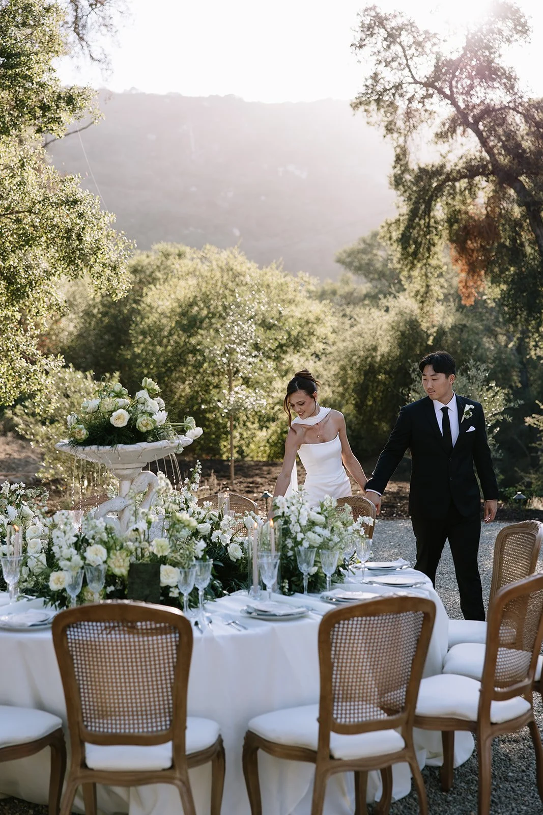 A couple dressed in wedding attire standing close together outdoors at sunset, holding a bouquet of flowers. The groom is in a mauve suit and the bride is in a white lace wedding dress.