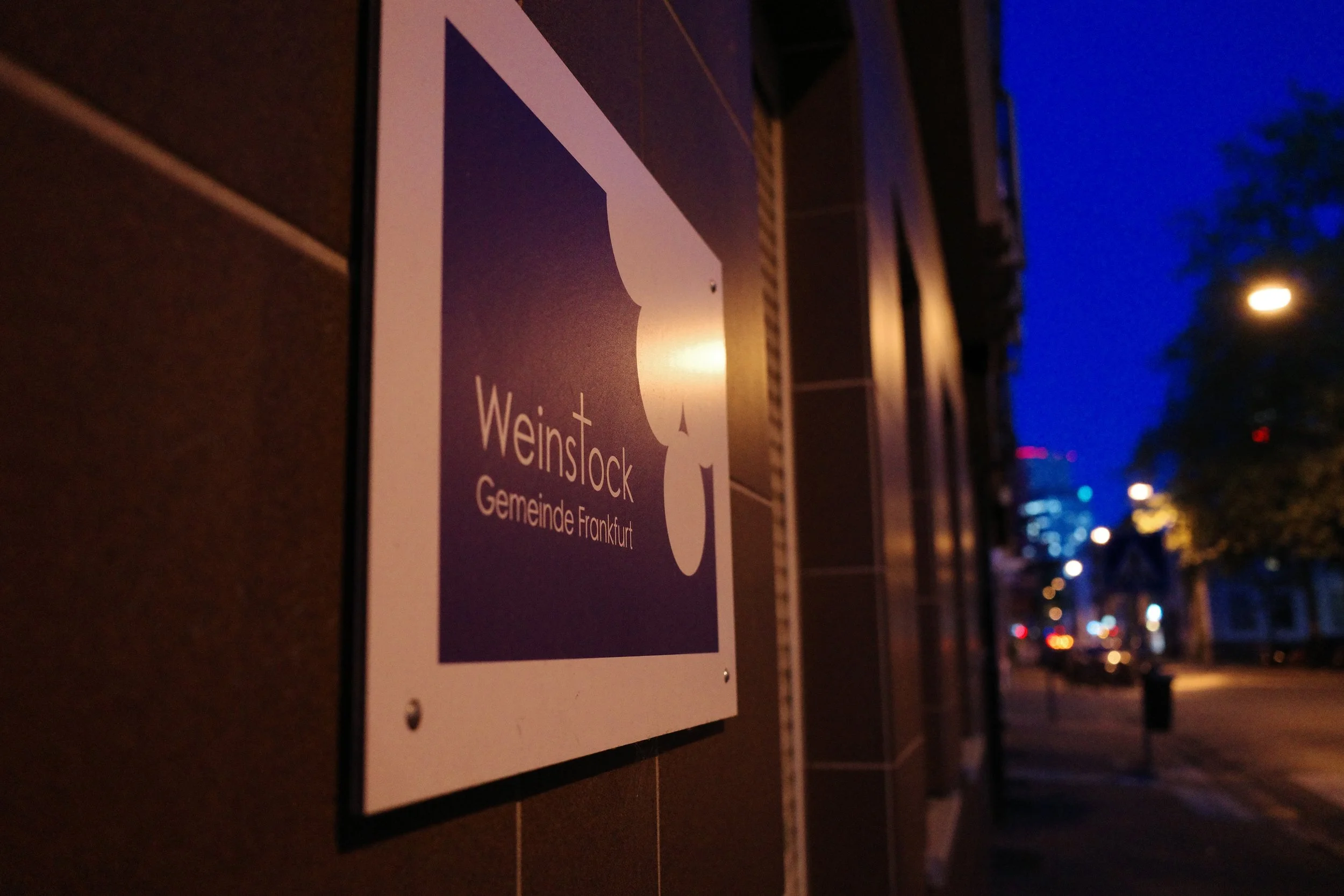 A close-up of a sign attached to a wall; the sign features a white logo and text that reads 'Weinstock Gemeinde Frankfurt' on a dark background. The image was taken outdoors at night, with streetlights and blurred city lights visible in the background.