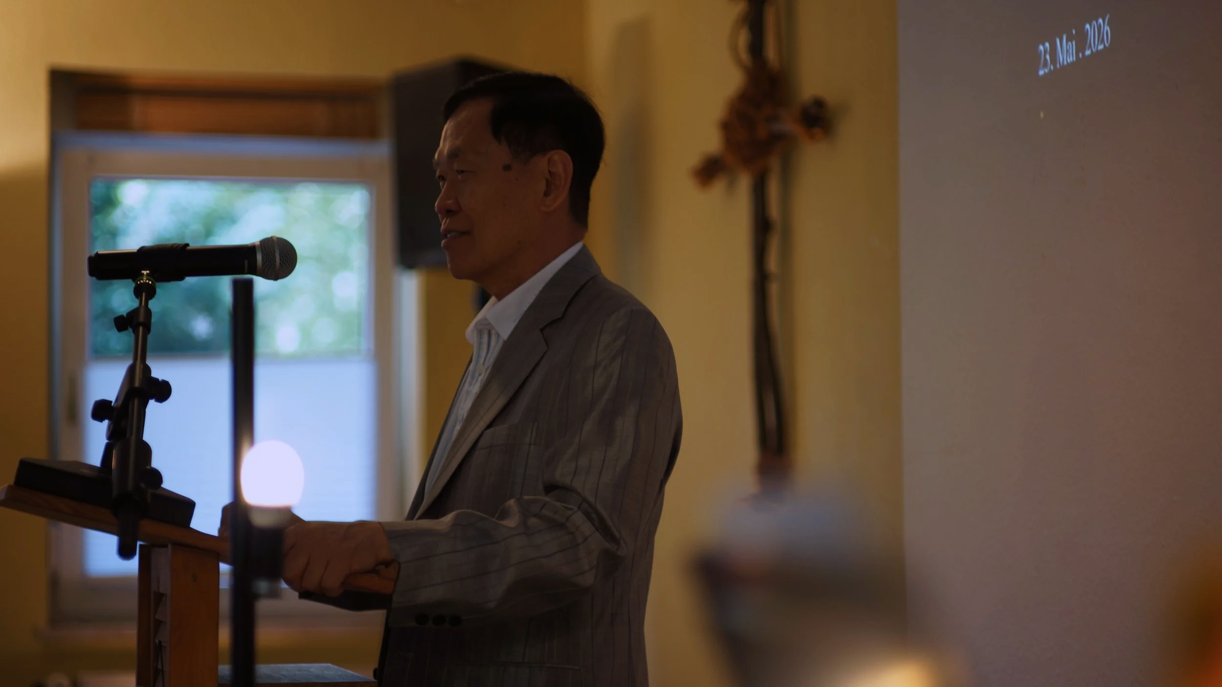 A man in a gray suit speaking at a podium with a microphone in a softly lit room with a window and a decorative cross on the wall. The image is dated May 23, 2026.