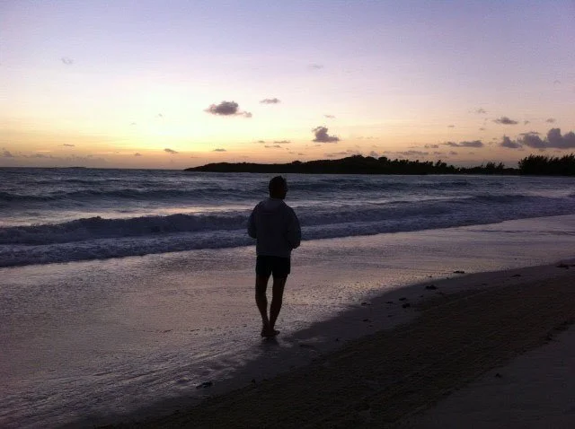 A person standing on the beach at sunset, looking at the ocean with gentle waves and a cloudy sky.