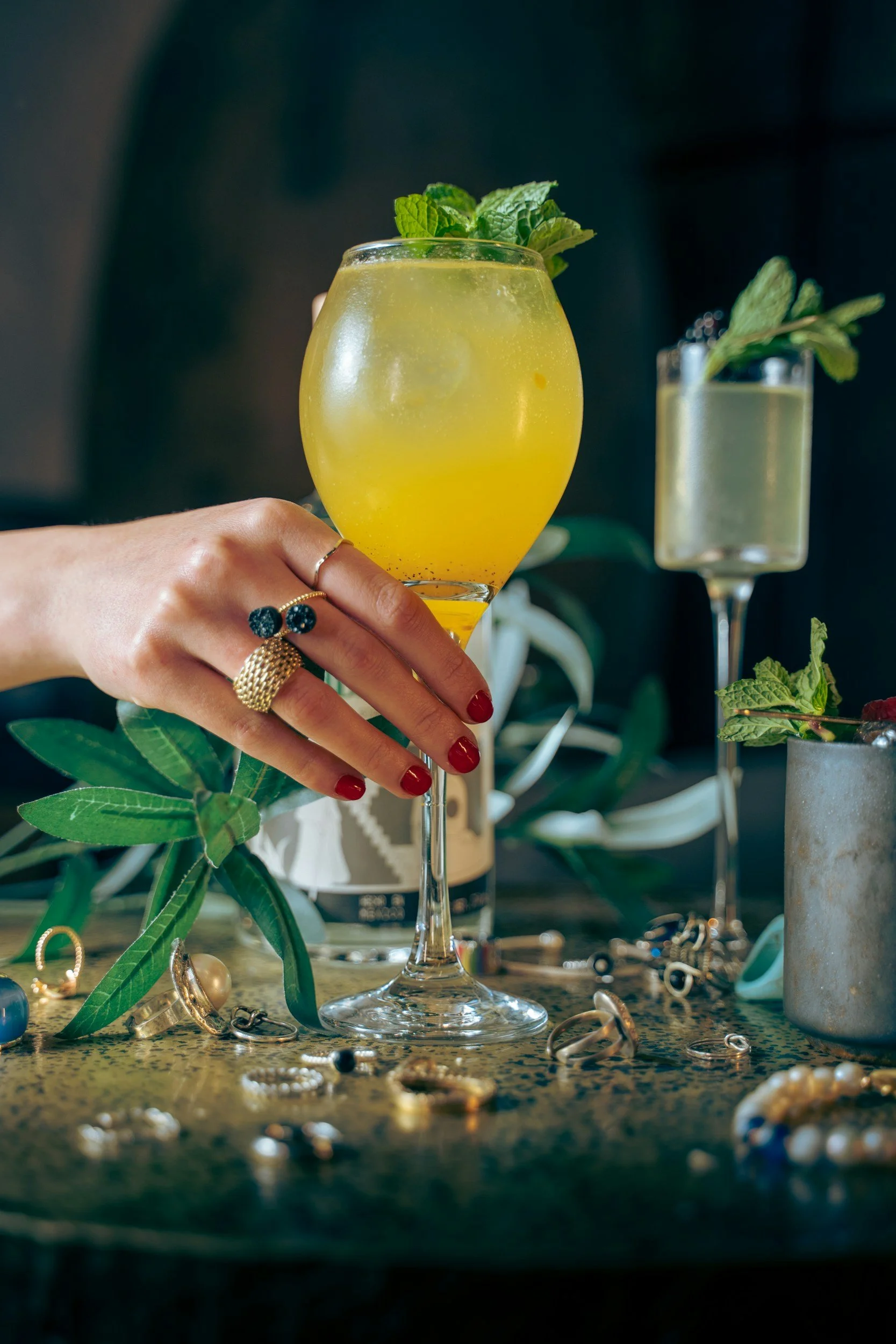 Hand with red-painted nails and gold rings holding a yellow cocktail garnished with mint leaves, surrounded by jewelry and other cocktails with mint garnish on a table.