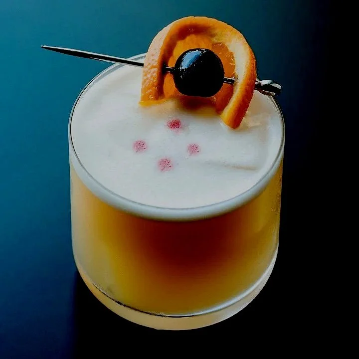 A creamy yellow cocktail with frothy top, garnished with an orange slice, a cherry on a skewer, and small pink edible dots.