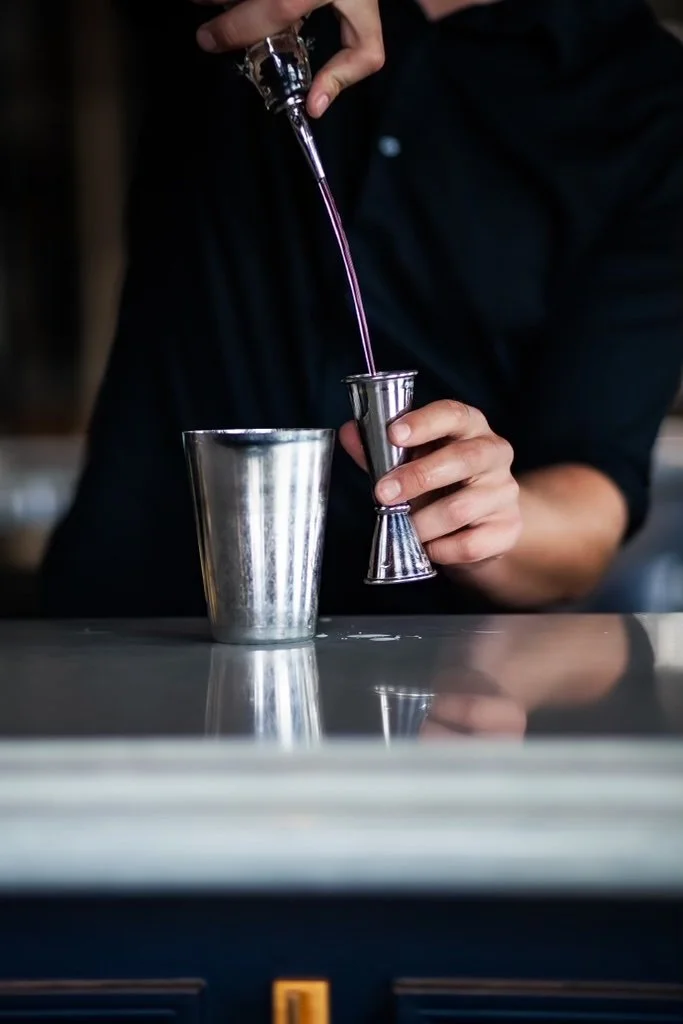 A bartender pouring purple liquid from a pour spout into a metal jigger.