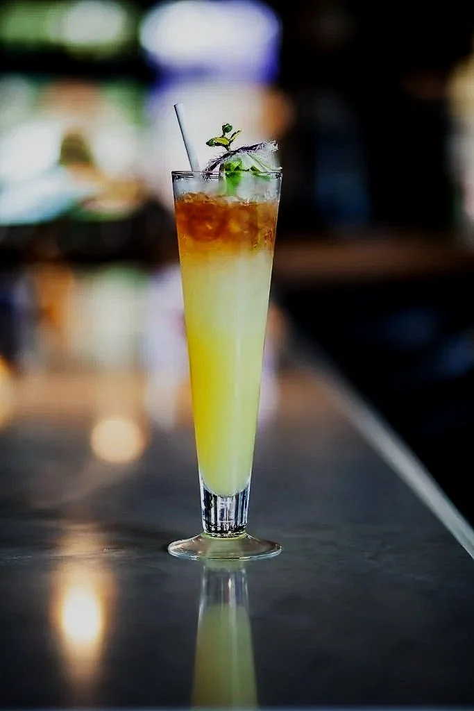 A tall glass cocktail with layered yellow and brownish colors, garnished with lime, mint, and a straw, on a bar counter.