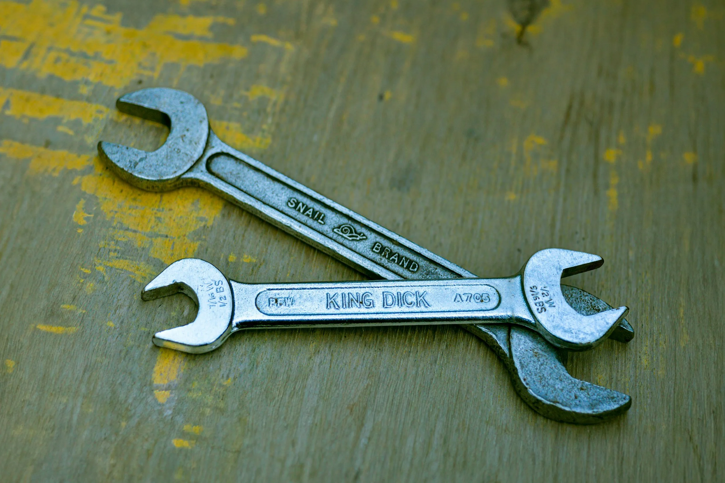 Two metal wrenches, one labeled "SNALL BRAND" and the other labeled "KING DICK," are crossed on a wooden surface with worn yellow paint.