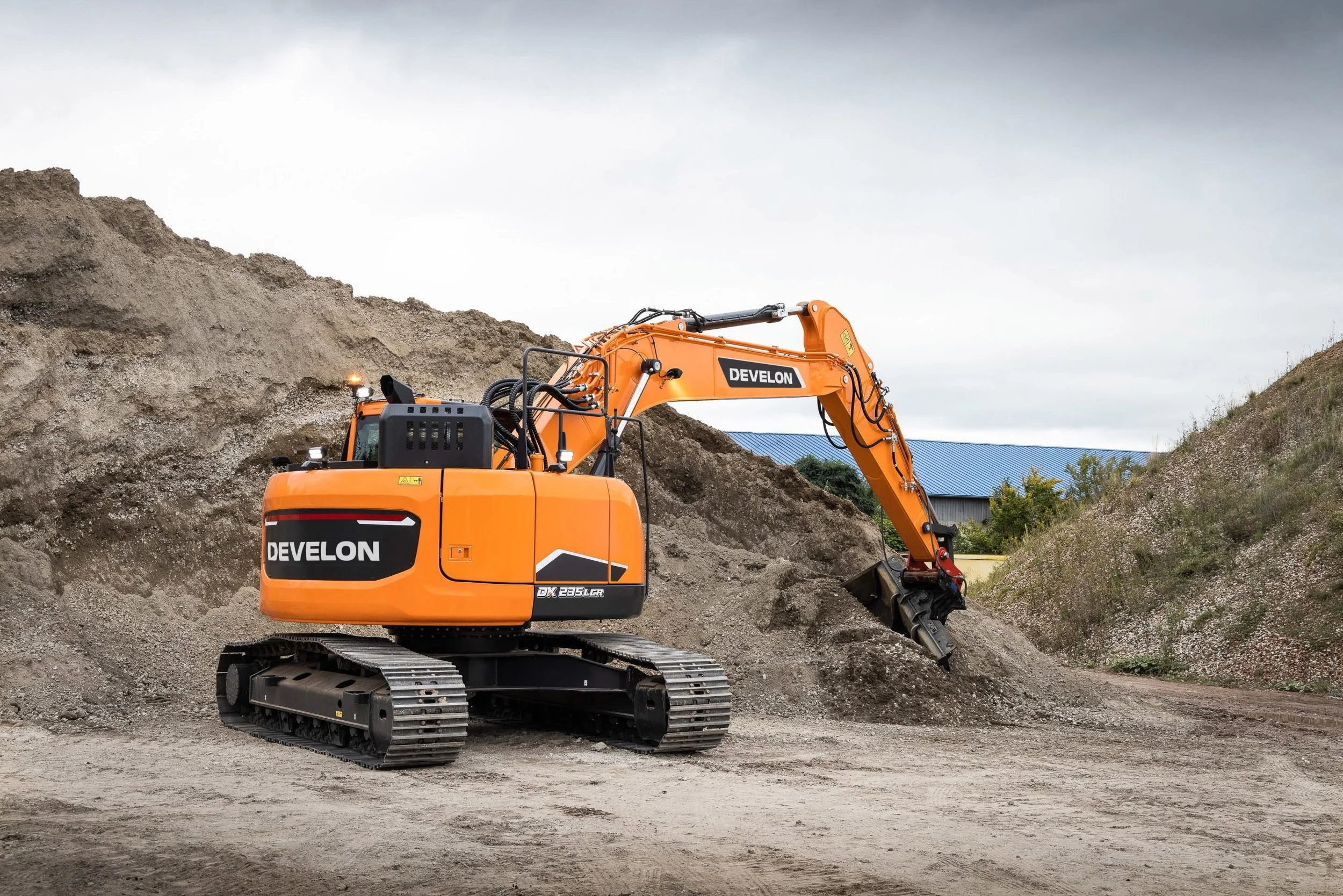 An orange Develon mini excavator working on a construction site, digging into a dirt hill with cloudy sky and a building with a blue roof in the background.