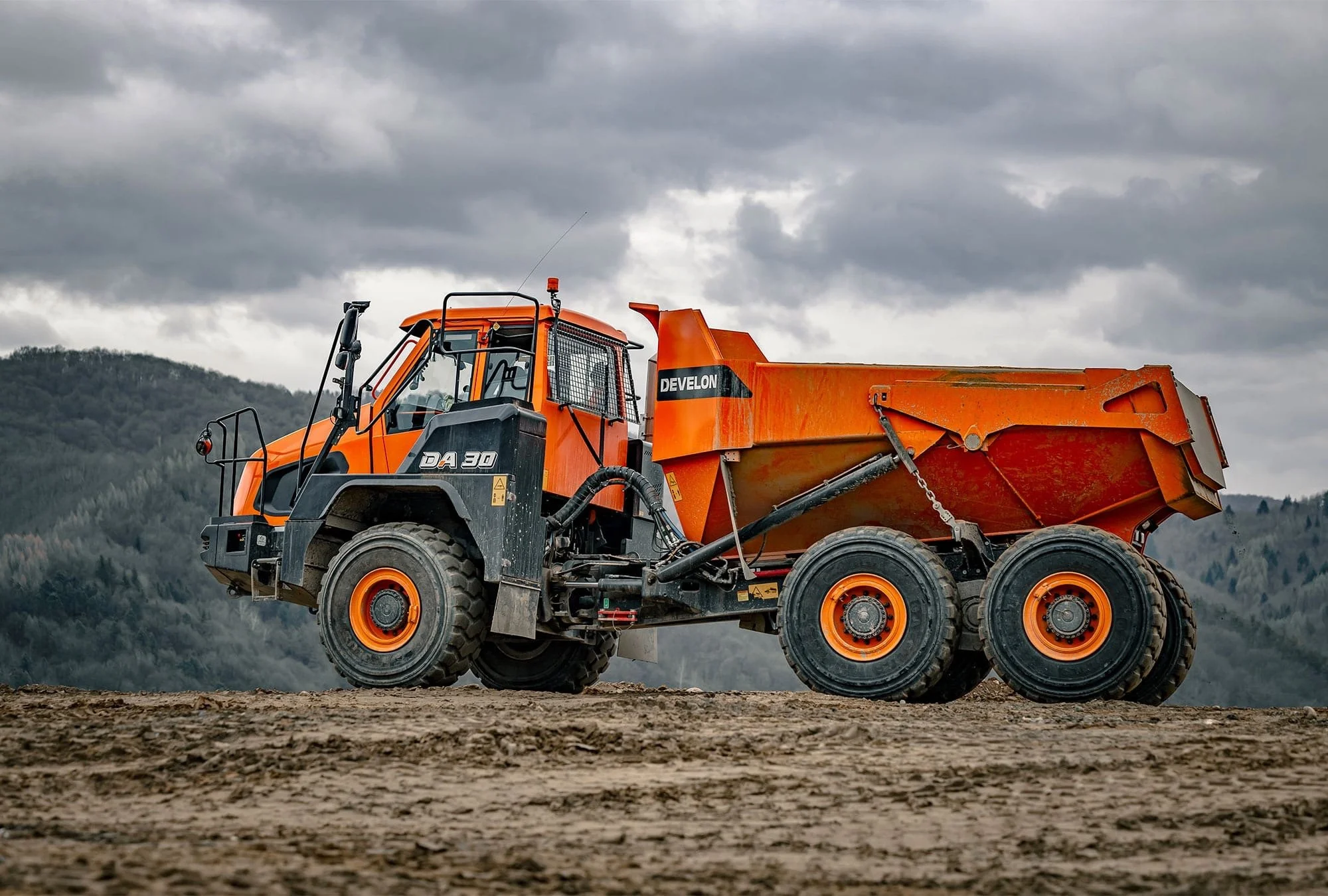 Orange heavy-duty dump truck on construction site, with a rugged landscape and cloudy sky in the background.