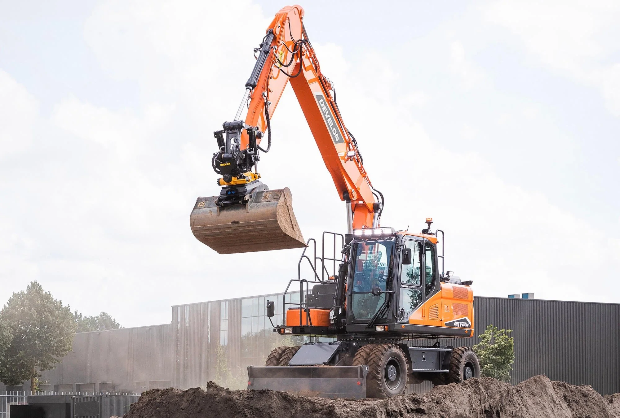 An orange Develon DX100W excavator working on a dirt hill, with large rugged tires, hydraulic arm, and a bucket attachment, outdoors on a cloudy day.