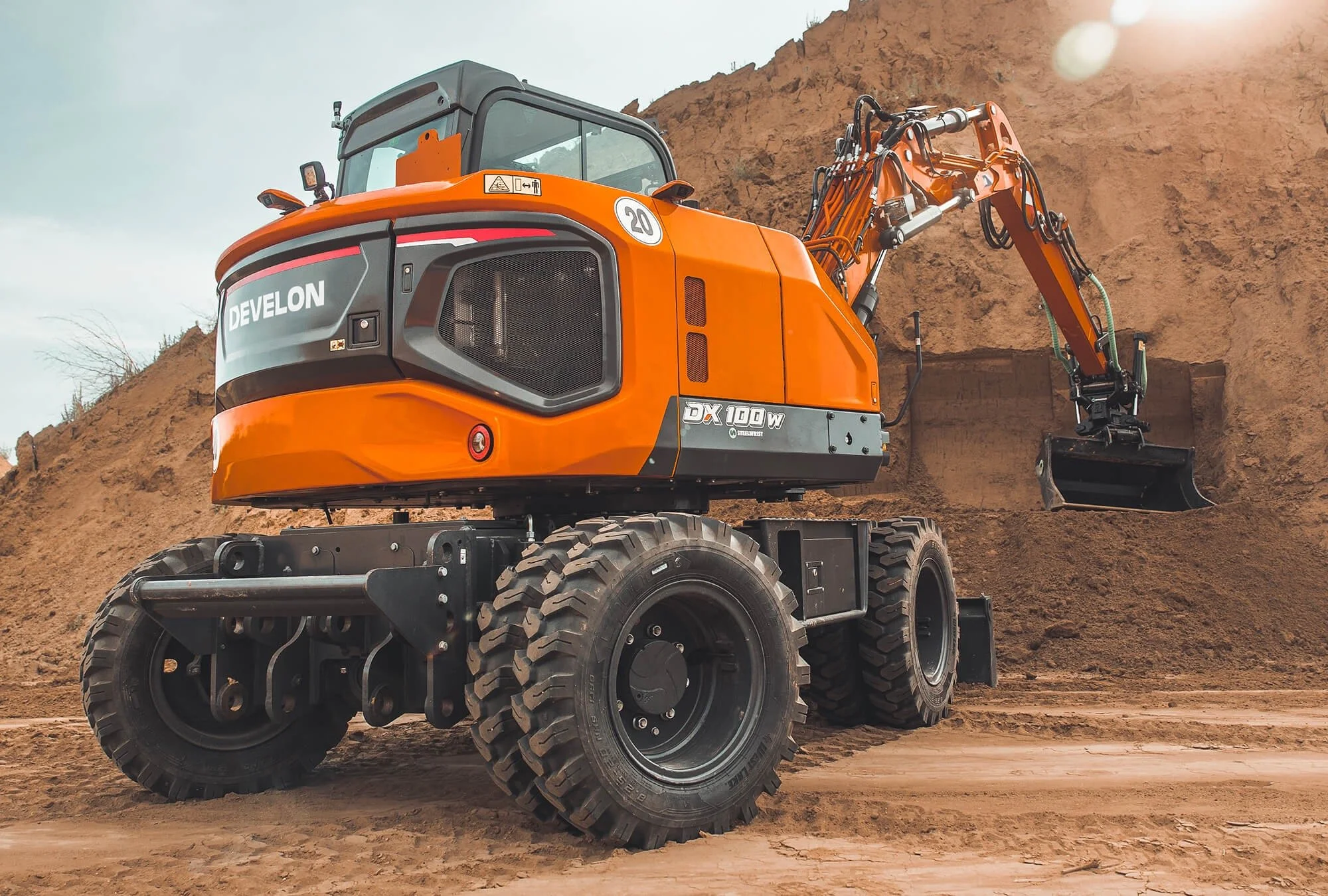 An orange Develon DX100W excavator working on a dirt hill, with large rugged tires, hydraulic arm, and a bucket attachment, outdoors on a cloudy day.
