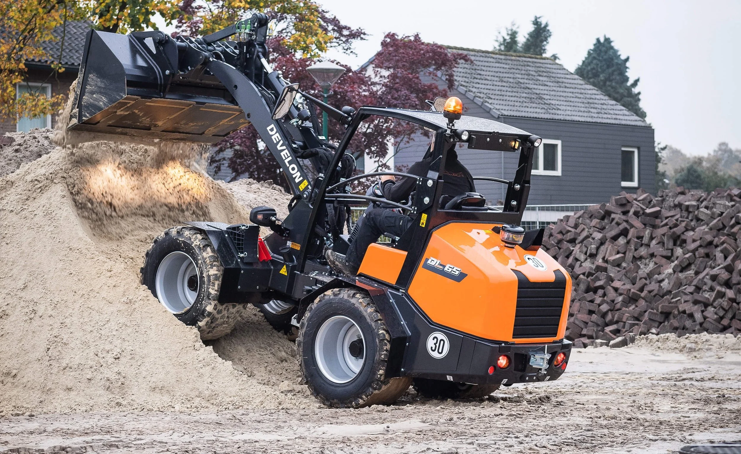 Orange compact excavator moving a pile of sand at a construction site, with a grey house and trees in the background.