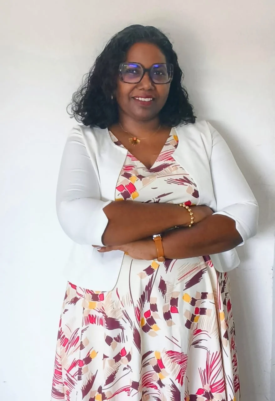 Femme confiante avec cheveux bouclés, porte des lunettes, un chemisier coloré et une veste blanche, posant avec les bras croisés contre un mur blanc.