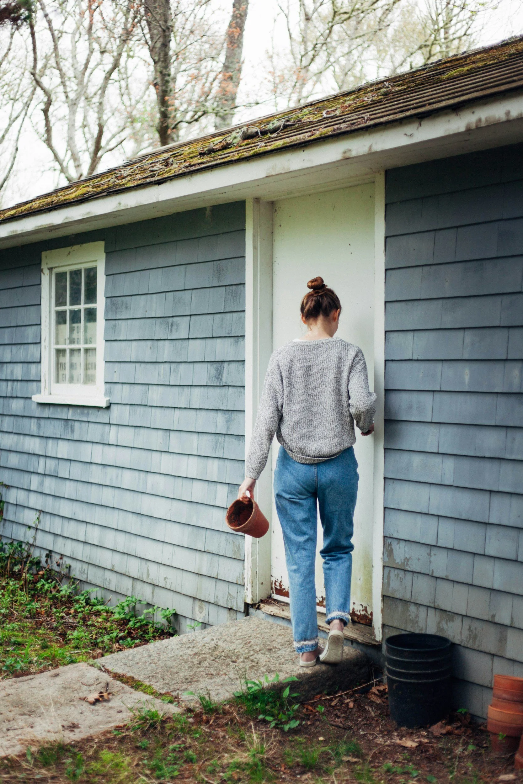 Woman walking toward a door of a small, blue house with gray shingles, holding a flowerpot, during daytime.