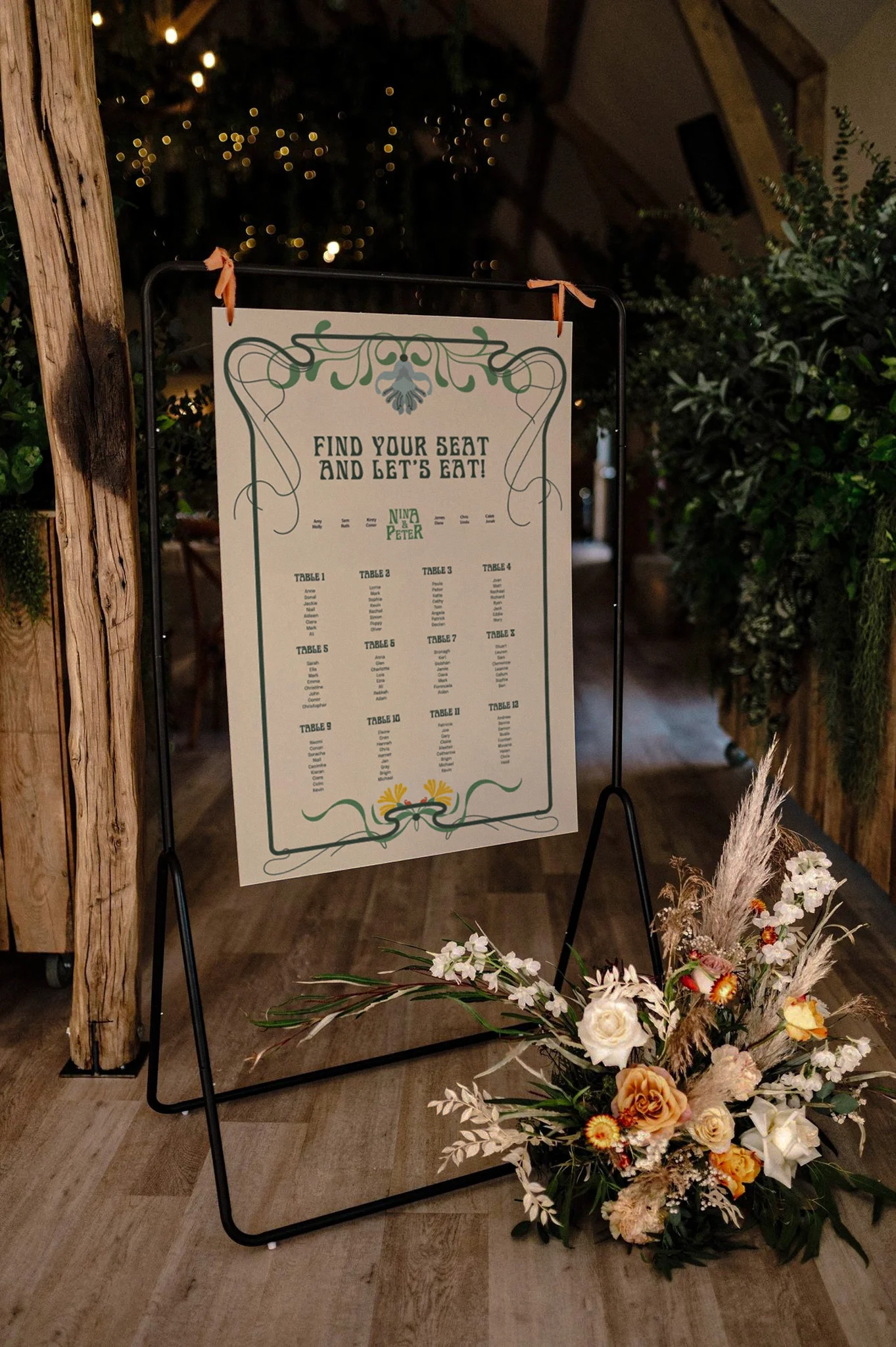 Wedding seating chart on a stand with a floral arrangement in front, decorated with an ornate border and the title "Find your seat and let's eat!" with names and table assignments.