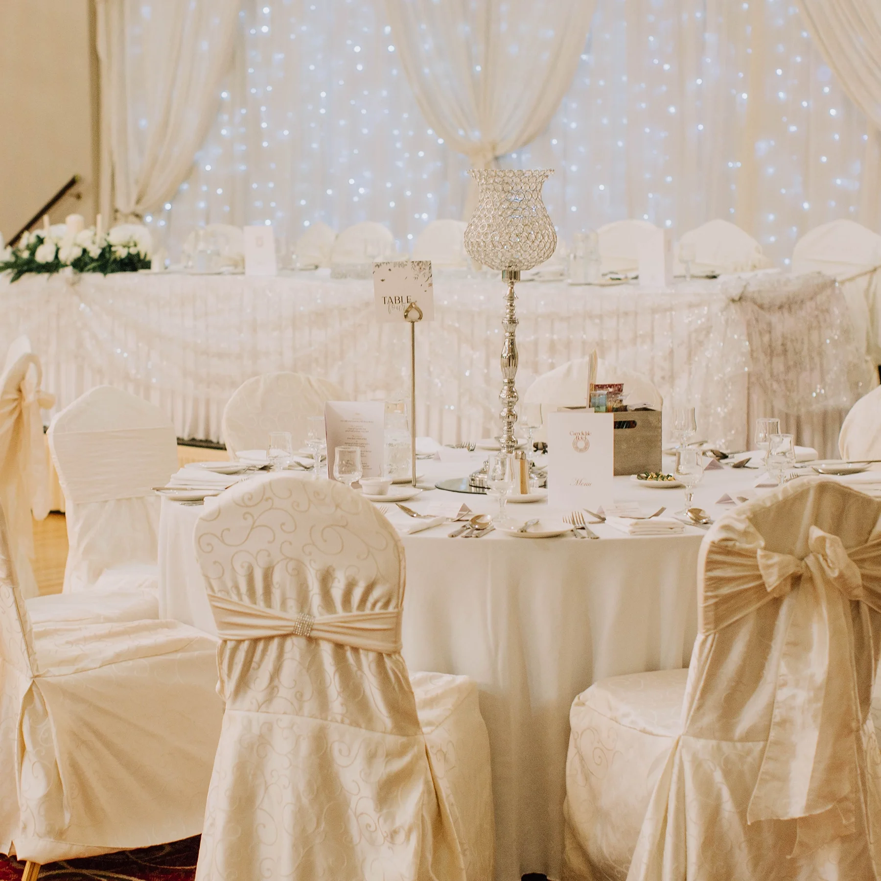 Elegant banquet table with white tablecloth, surrounded by white chairs with satin covers and bows. Decor includes a tall crystal candleholder and a curtain backdrop with twinkling lights.