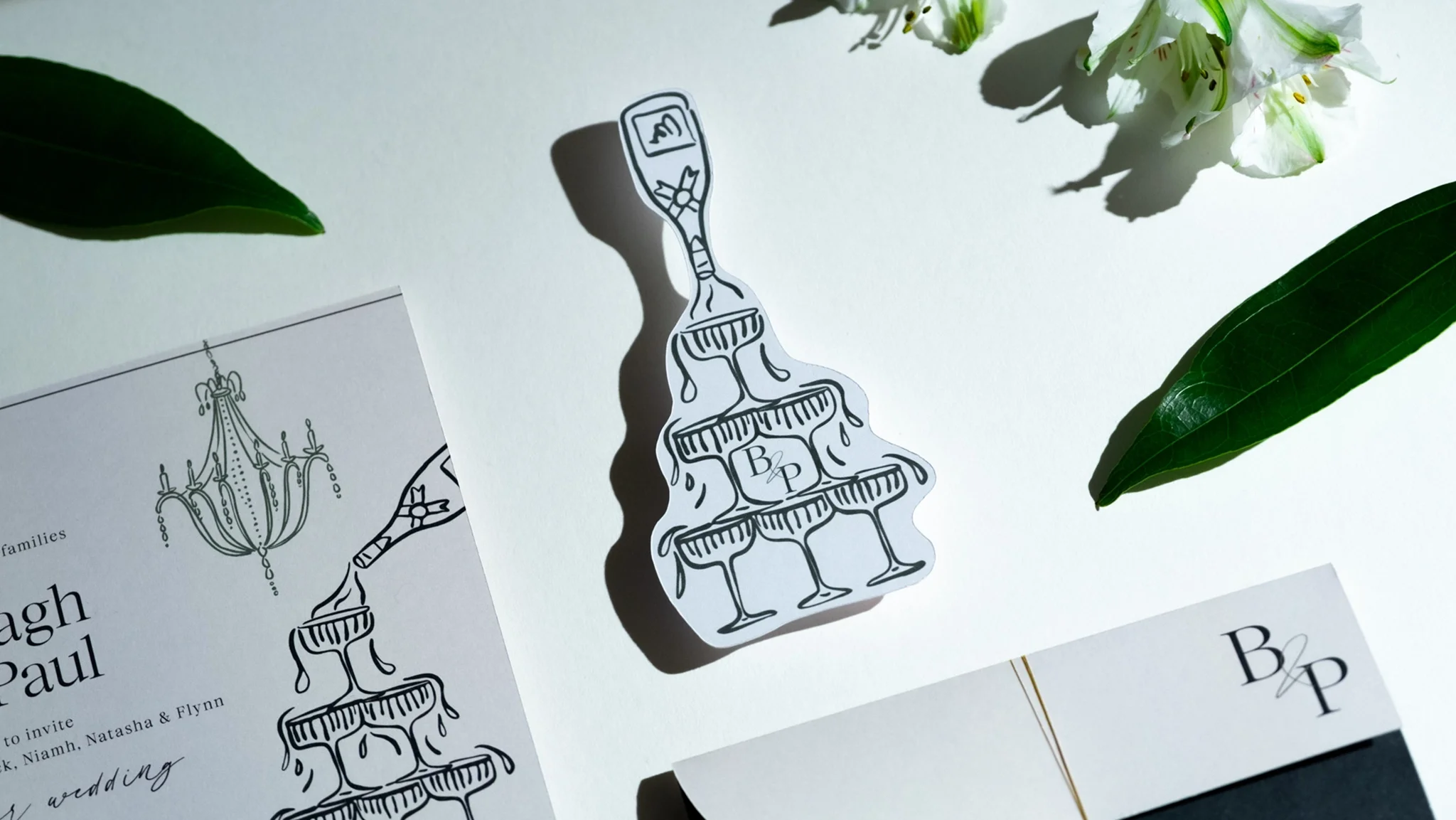Invitation card with a black and white illustration of a champagne tower, decorated with the initials "B & P" on the bottom glass, accompanied by green leaves and white flowers.