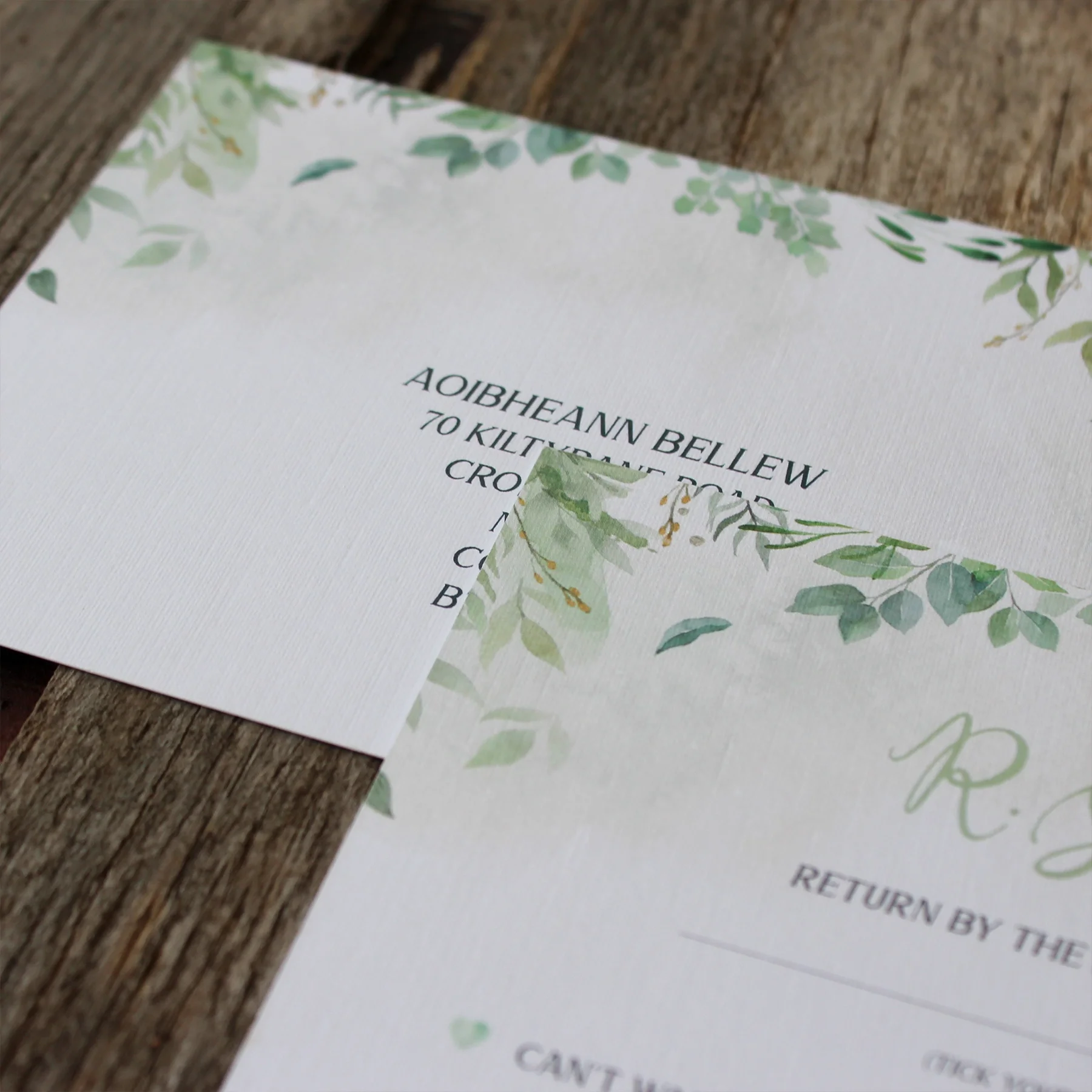 Close-up of wedding invitation with green foliage border on white textured paper placed on wooden surface.