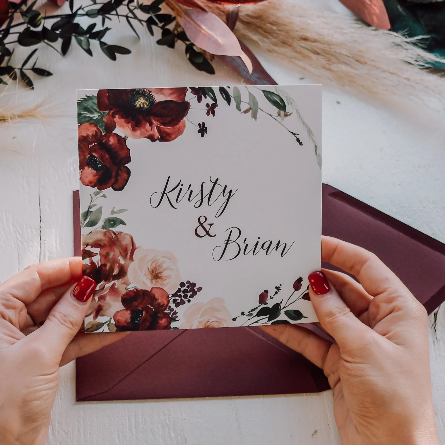 A person with red painted nails holding a wedding invitation card with floral design and the names Kirsty & Brian.