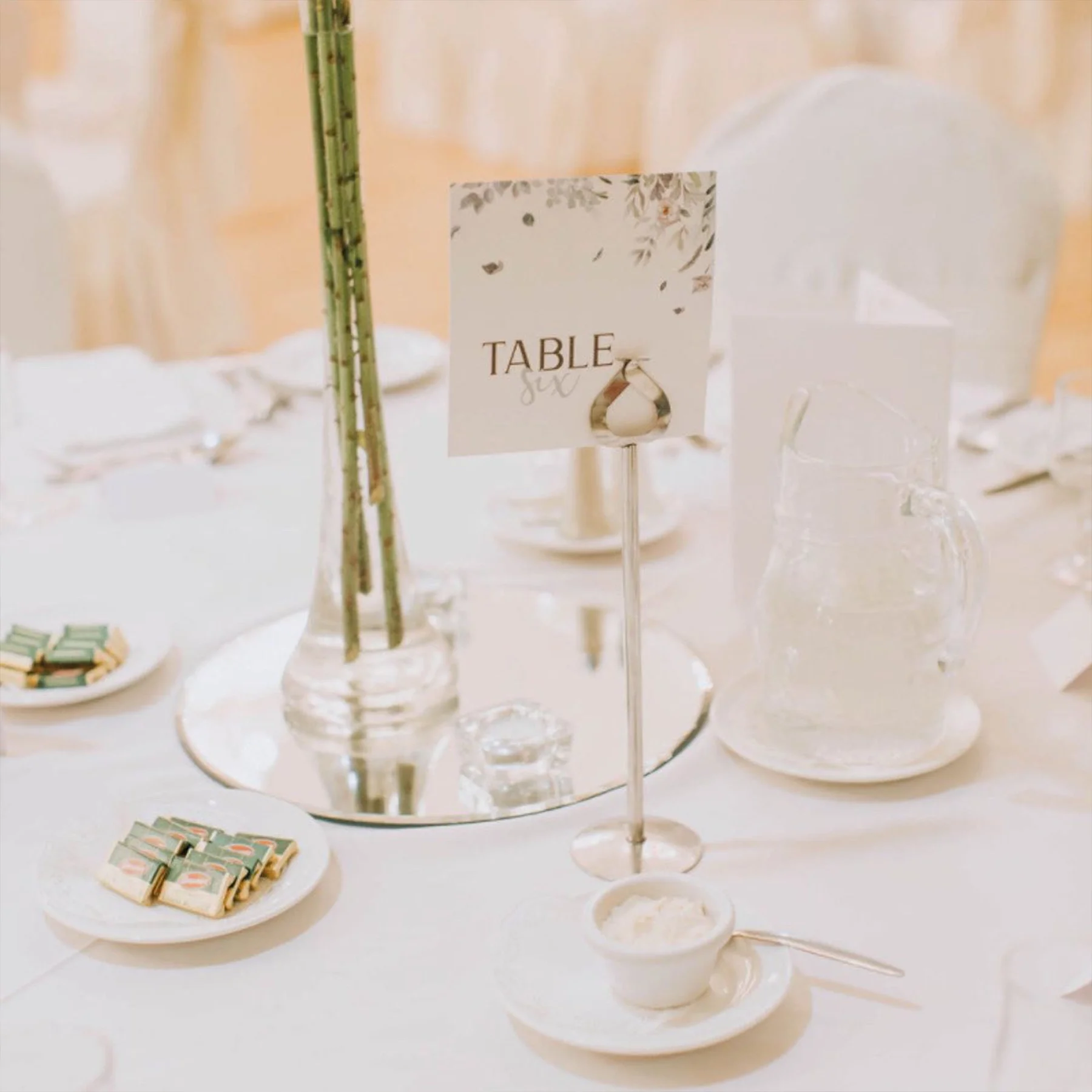 Elegant wedding table setting with white tablecloth, floral centerpiece, and a table sign. Clear glass pitcher and small bowl of cream or dip placed on a white dish. Plates with small packages or favors and glassware scattered around.