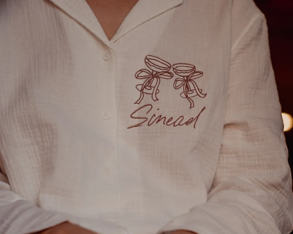 A person wearing a cream-colored button-up shirt with embroidered two tied ribbon bows and the word 'Sinead' below it.