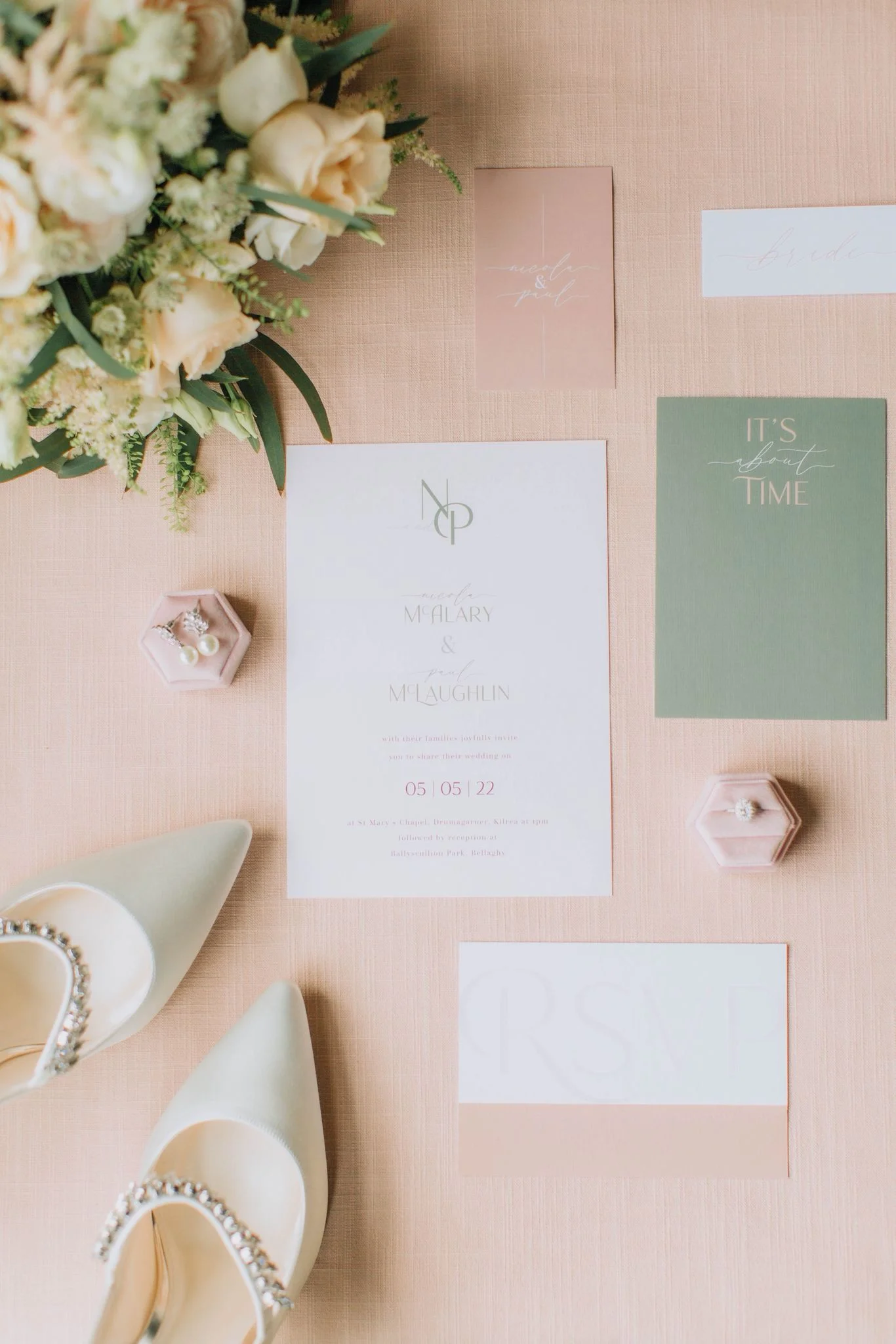 Wedding invitation flat lay with pink and green accents, pearl earrings, wedding shoes with rhinestone details, and a bouquet of flowers in the top left corner.