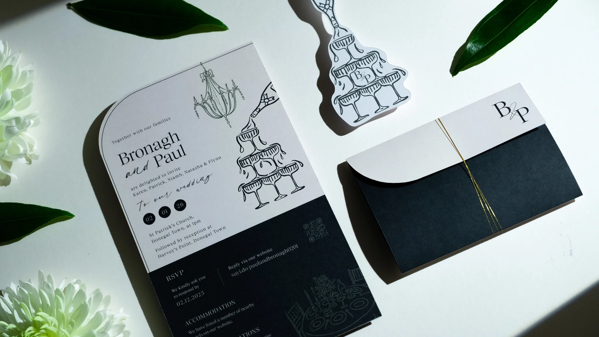 Wedding invitation card with black and white design, featuring an illustration of a layered champagne tower, accompanied by an envelope with black and white design elements, placed on a white surface with green leaves and white flowers.