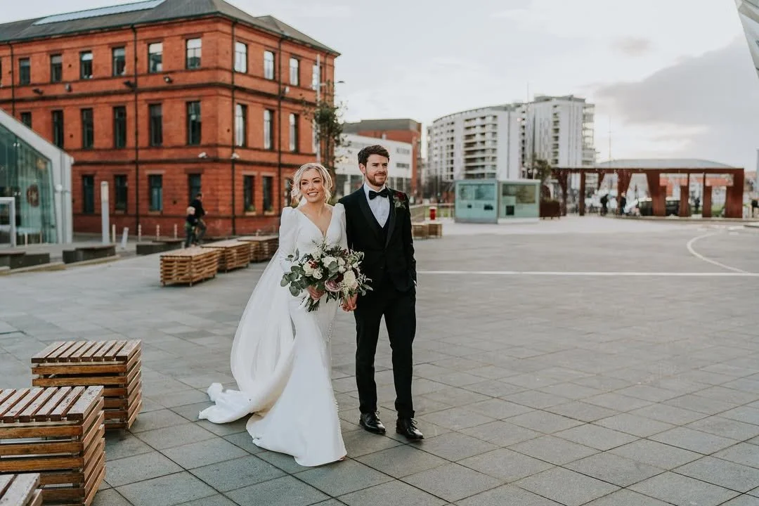A bride and groom walking outdoors in a city square, with the bride holding a bouquet of flowers, smiling and dressed in a white gown, and the groom in a black tuxedo with a bow tie, surrounded by modern buildings and benches.