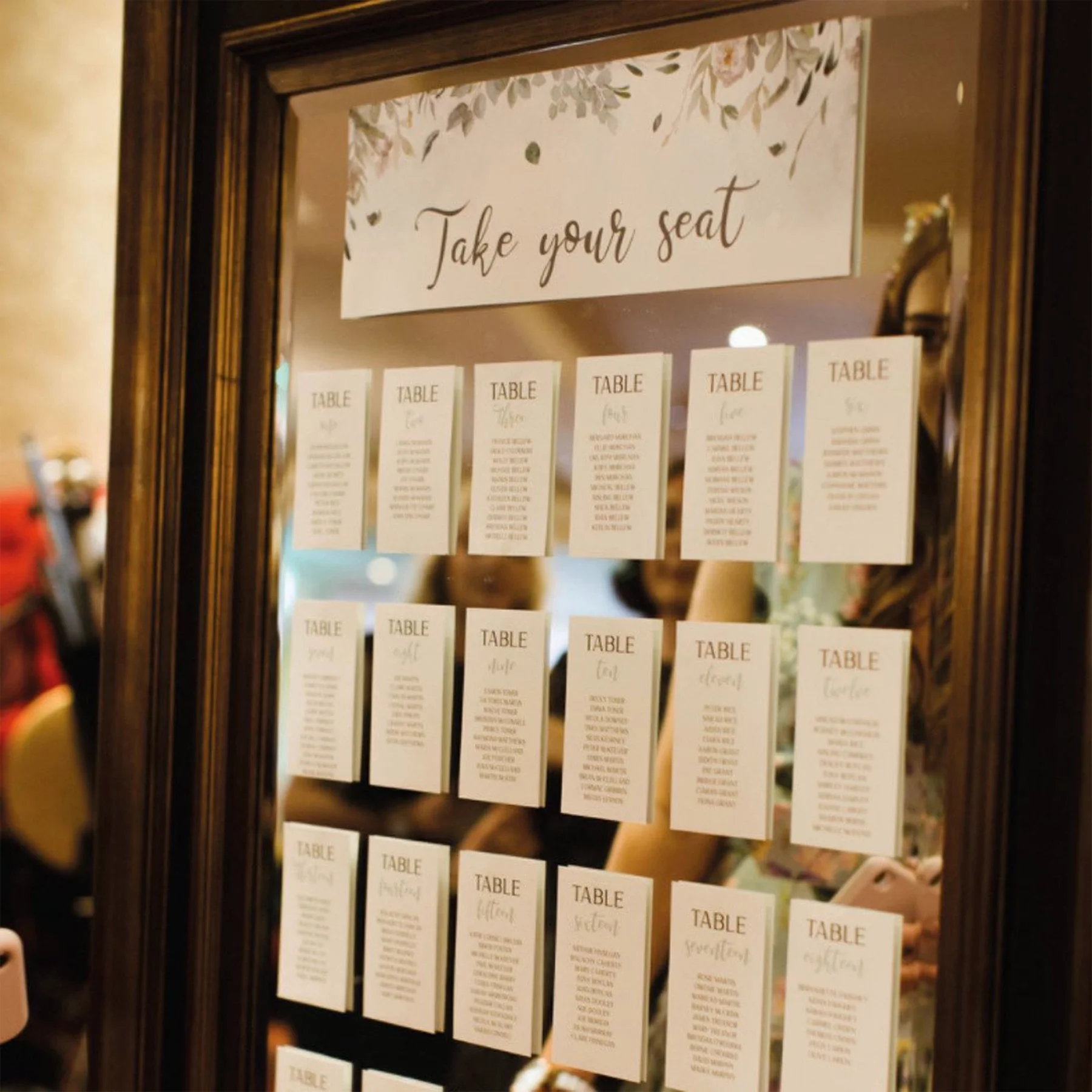 A mirror with a sign that says 'Take your seat' at the top, and various table number cards below, hung in a grid pattern.