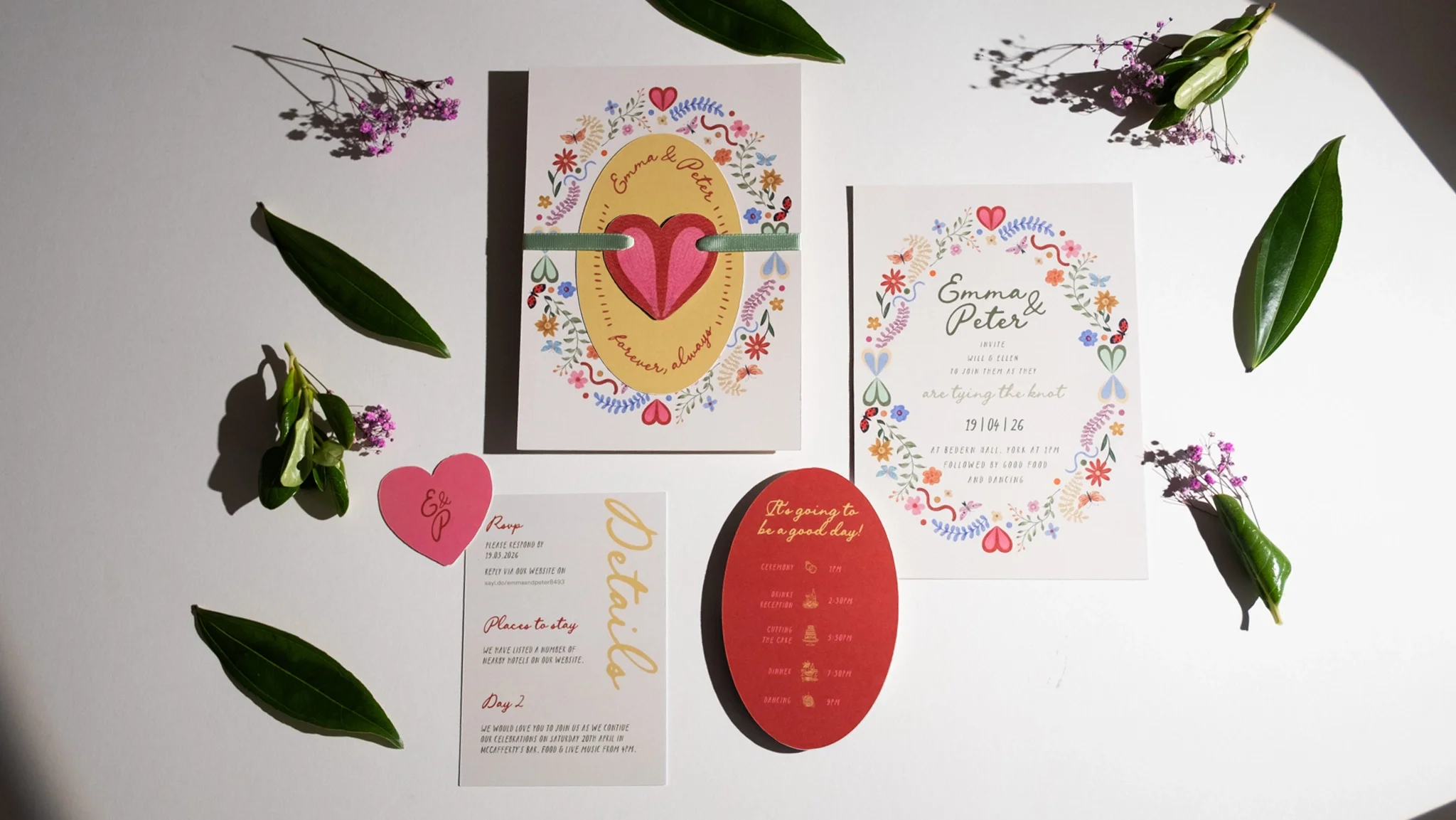 Wedding invitations with floral designs, pink hearts, and event details, surrounded by green leaves and small purple flowers on a white surface.