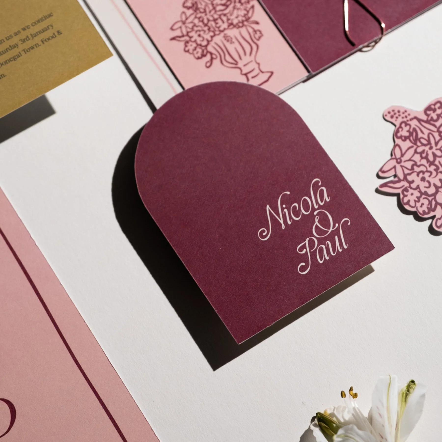 A burgundy wedding invitation with the names "Nicola & Paul" in white cursive font, surrounded by decorative floral illustrations and background papers in pink, white, and gold.