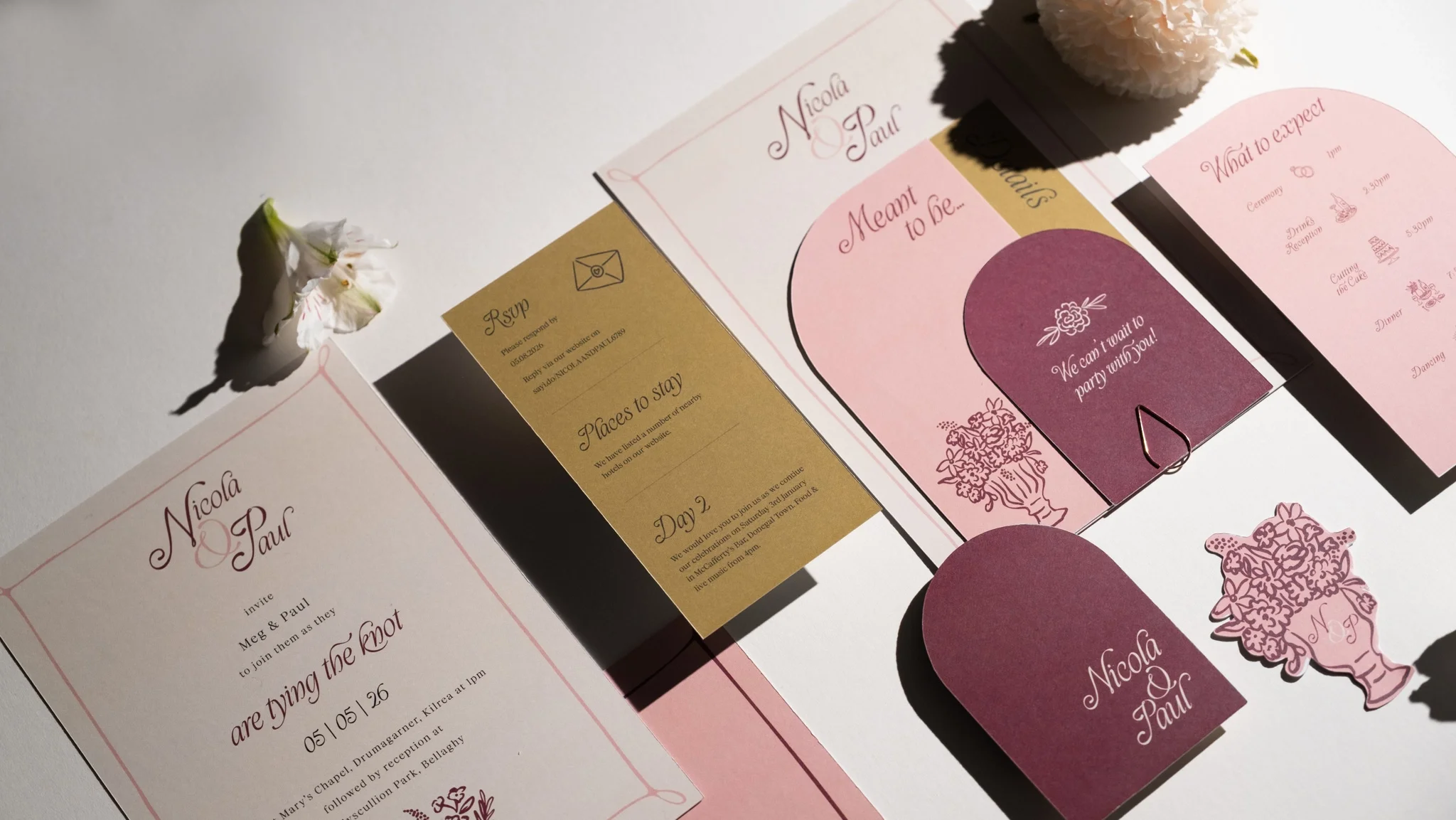 Wedding invitation and related paper items on a white surface, decorated with small flowers and cutouts. The invitation has pink and burgundy colors with cursive writing, mentions Nicola and Paul, and includes details about the event.
