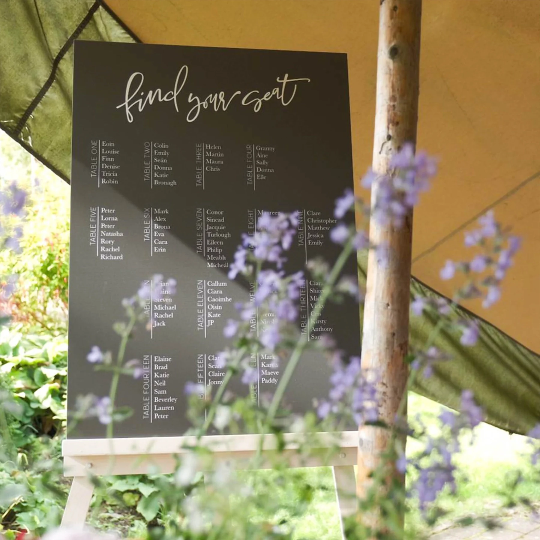 A seating chart for a wedding or event, titled 'find your seat,' with tables numbered One to Fifteen, each listing names of guests. The chart is black with white text, setup on an easel, and partially obscured by flowering plants in the foreground.