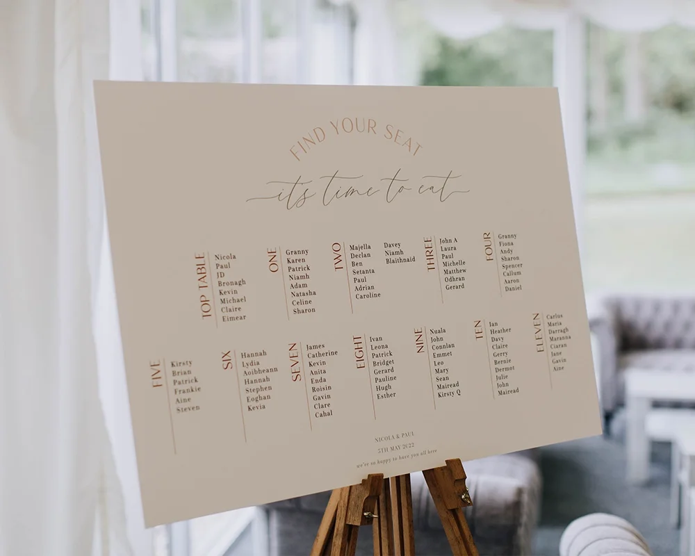A wedding seating chart with a cream background and elegant text, displayed on an easel. The chart lists tables numbered one to eleven with guest names.
