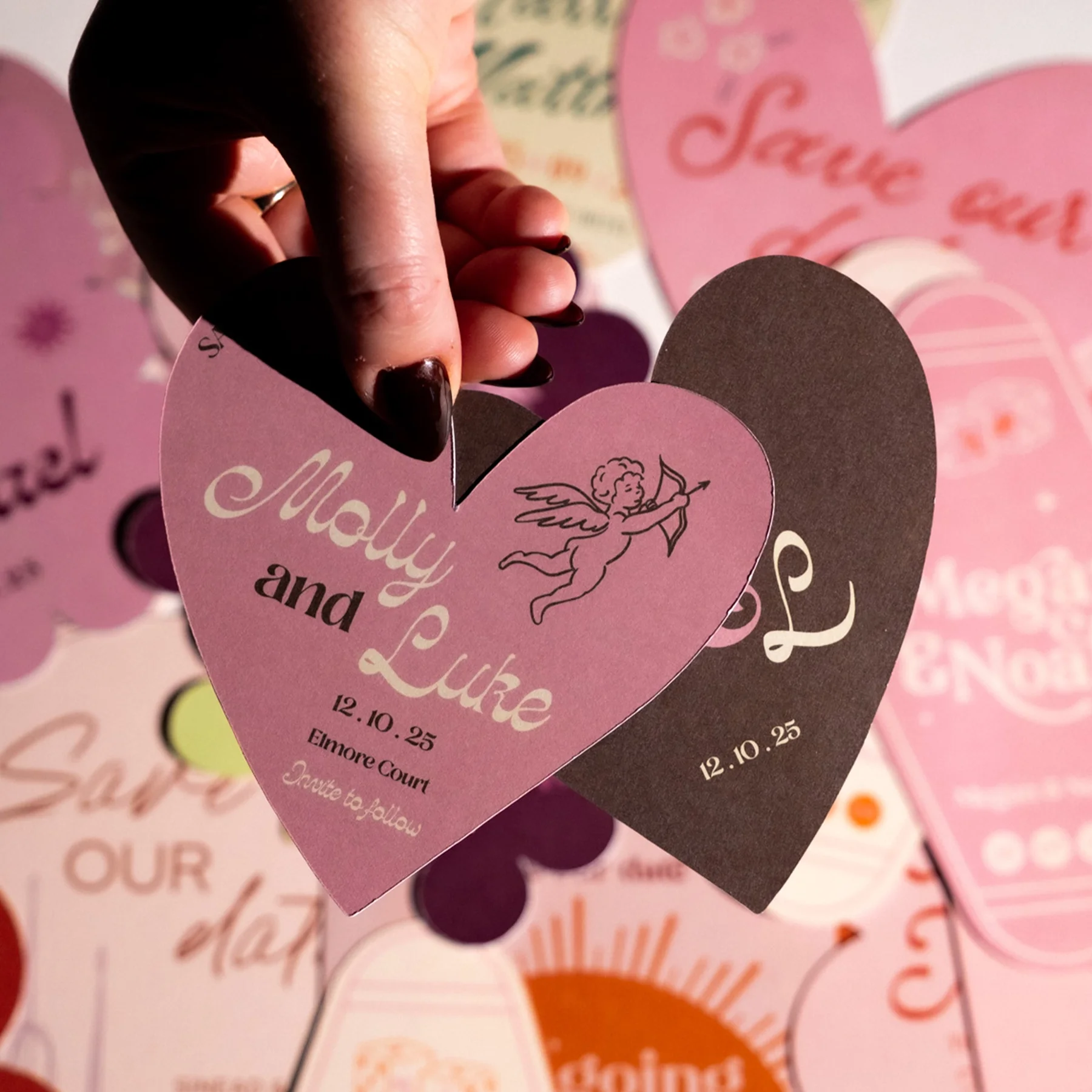 A hand holding two overlapping wedding invitations, one pink and one brown, with a cherub holding a bow and arrow illustration. The pink invitation reads 'Molly and Luke', with the date '12.10.25' and location 'Elmore Court'. The background features 