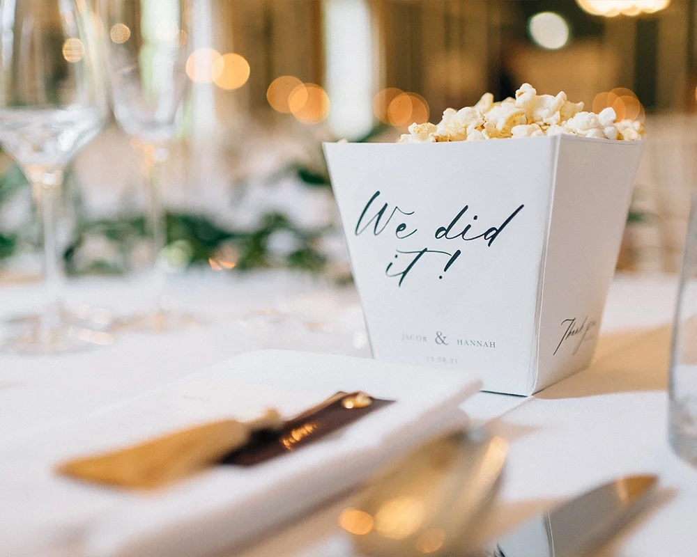 A wedding reception table with a white popcorn bucket labeled "We did it!" in elegant script, alongside wine glasses and table settings with soft lighting in the background.