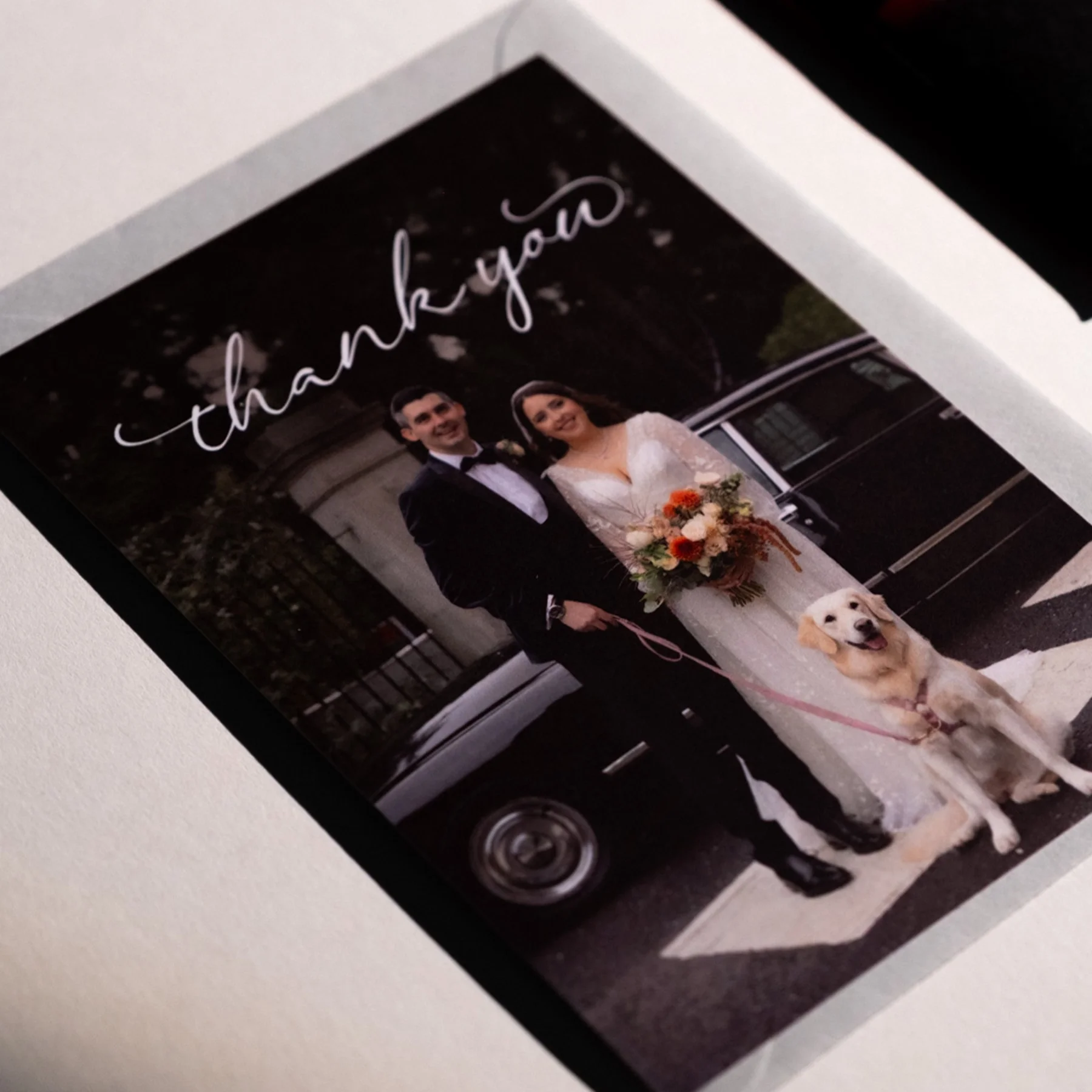Wedding photo of a bride and groom standing outdoors next to a black car, with a dog sitting in front of them. The bride is holding a bouquet, and the words 'thank you' are written at the top of the photo.
