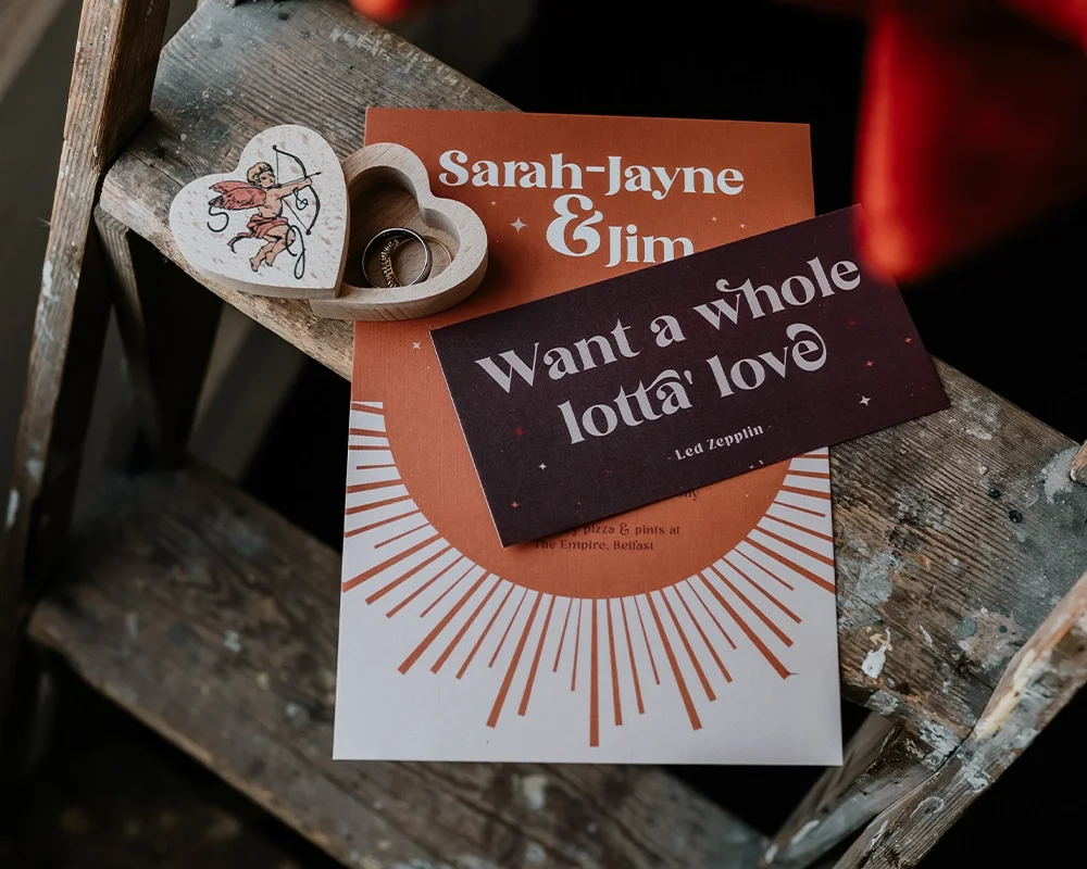 A wooden heart-shaped box with two wedding rings, and two cards on a rustic wooden surface. One card reads 'Sarah-Jayne & Jim,' and the other is a quote by Lea Zeppin: 'Want a whole lotta love.'
