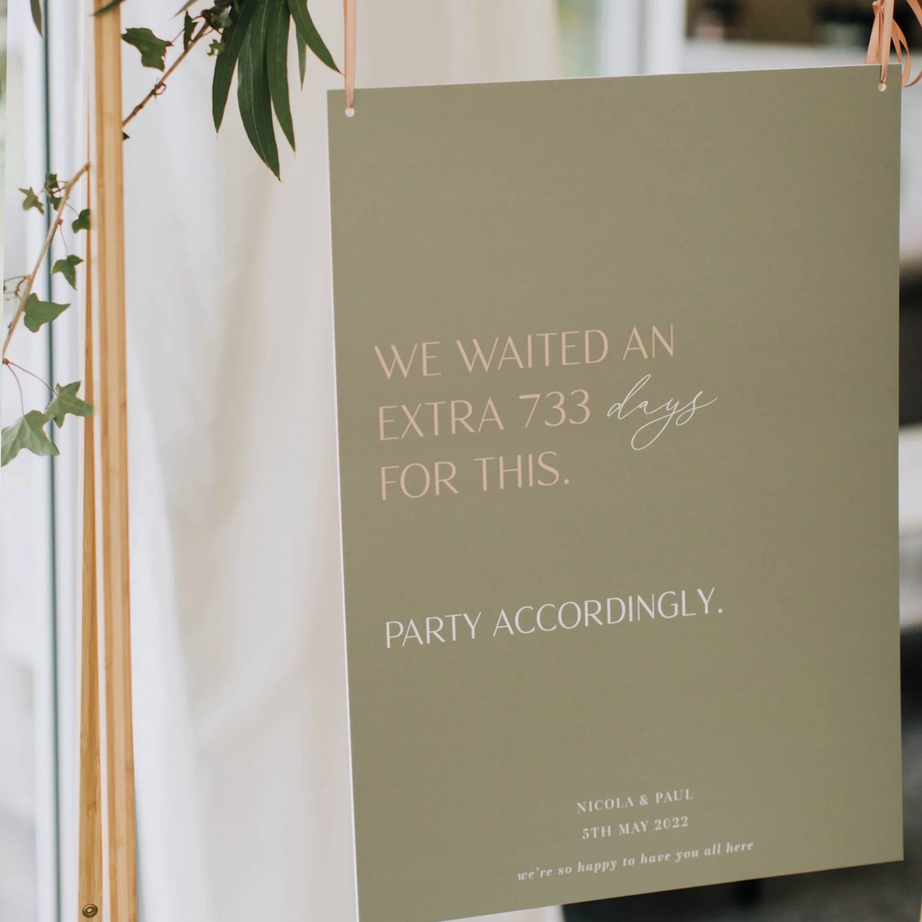 A green wedding sign hanging from a wooden stand with green leaves in the background, displaying in white and gray text: "We waited an extra 733 days for this. Party accordingly. Nicola & Paul, 5th May 2022, we're so happy to have you all here."
