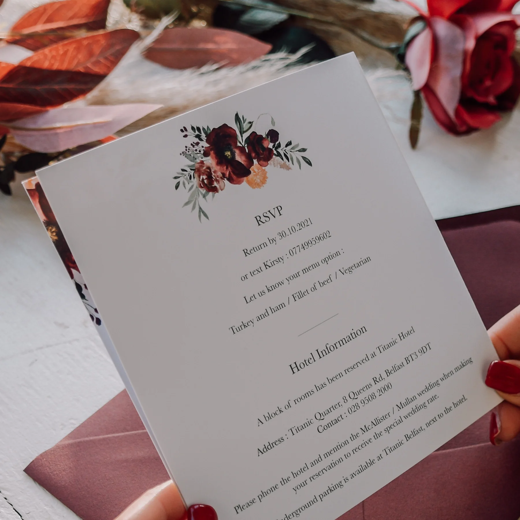 Wedding RSVP card featuring floral design at the top, with event details and hotel information printed below, held by a person with red painted nails.