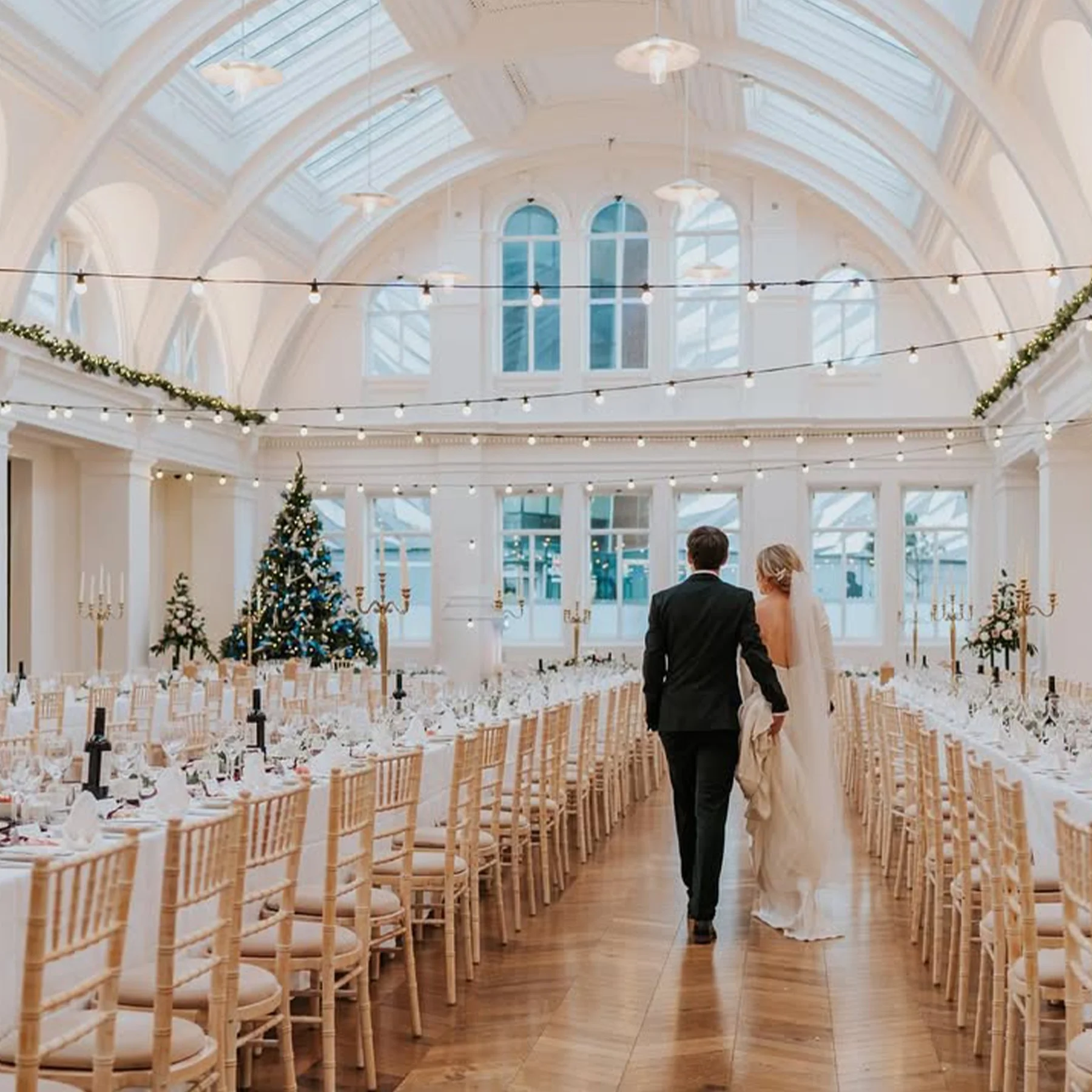A bride and groom walking through a decorated wedding reception hall with a Christmas tree, candles, and string lights.