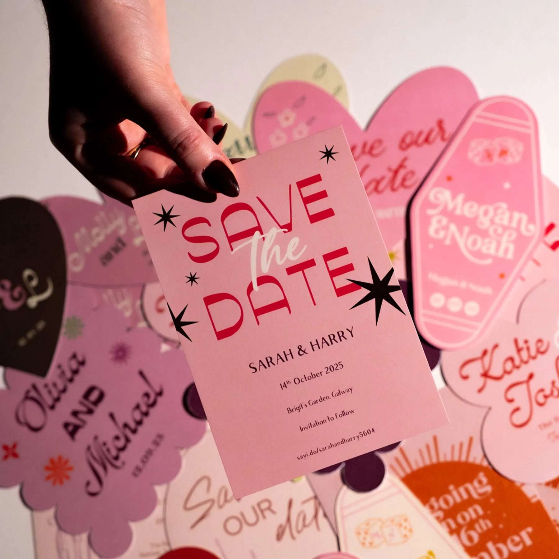 Pink save the date card for Sarah and Harry's wedding, dated October 14, 2025, with decorative stars and floral elements, held by a person with dark nail polish, against a background of various colorful wedding invitations and cards.