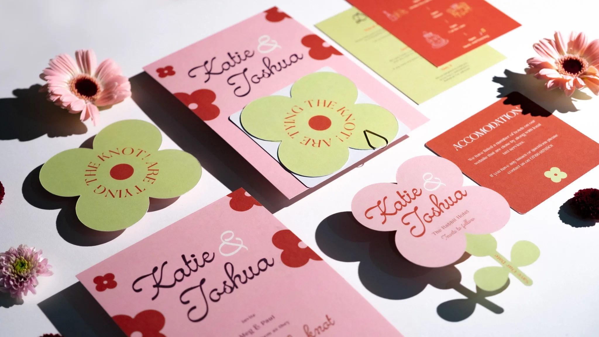 Flat lay photography of a modern pink and green wedding stationery suite for a happy couple, featuring 70s-inspired retro floral designs, die-cut flower RSVP cards, and colourful typography.